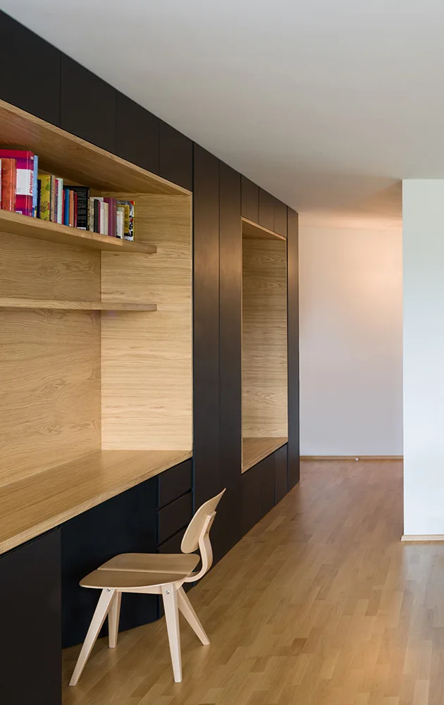 A minimalist home office featuring a matte black wall unit with an integrated light wood desk niche and a matching wooden chair.