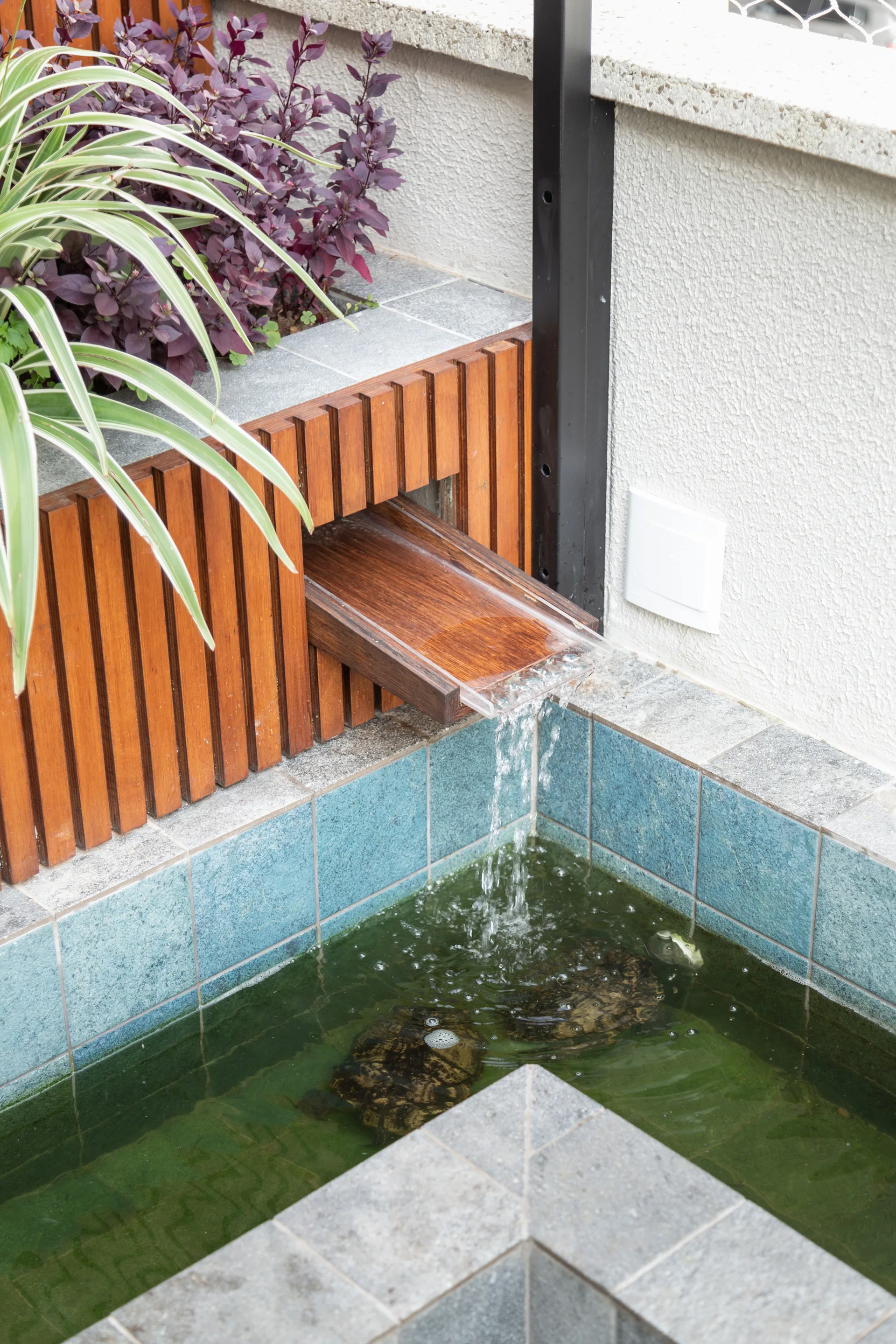 A small outdoor pond with blue tiles and swimming turtles features water pouring from a wooden spout built into a slatted wood planter.