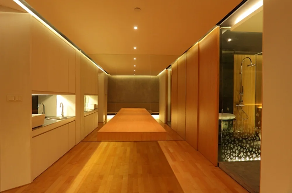 A minimalist corridor featuring a long floating wooden table, built-in kitchen cabinets with a sink, and a glass shower stall.