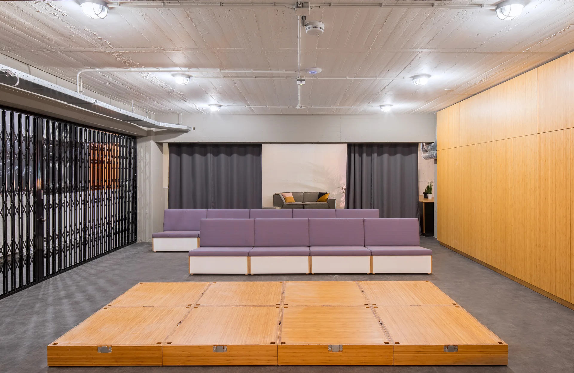 A multi-purpose industrial room features modular purple benches, a low raised bamboo platform, a wooden wall panel, and an exposed concrete ceiling.