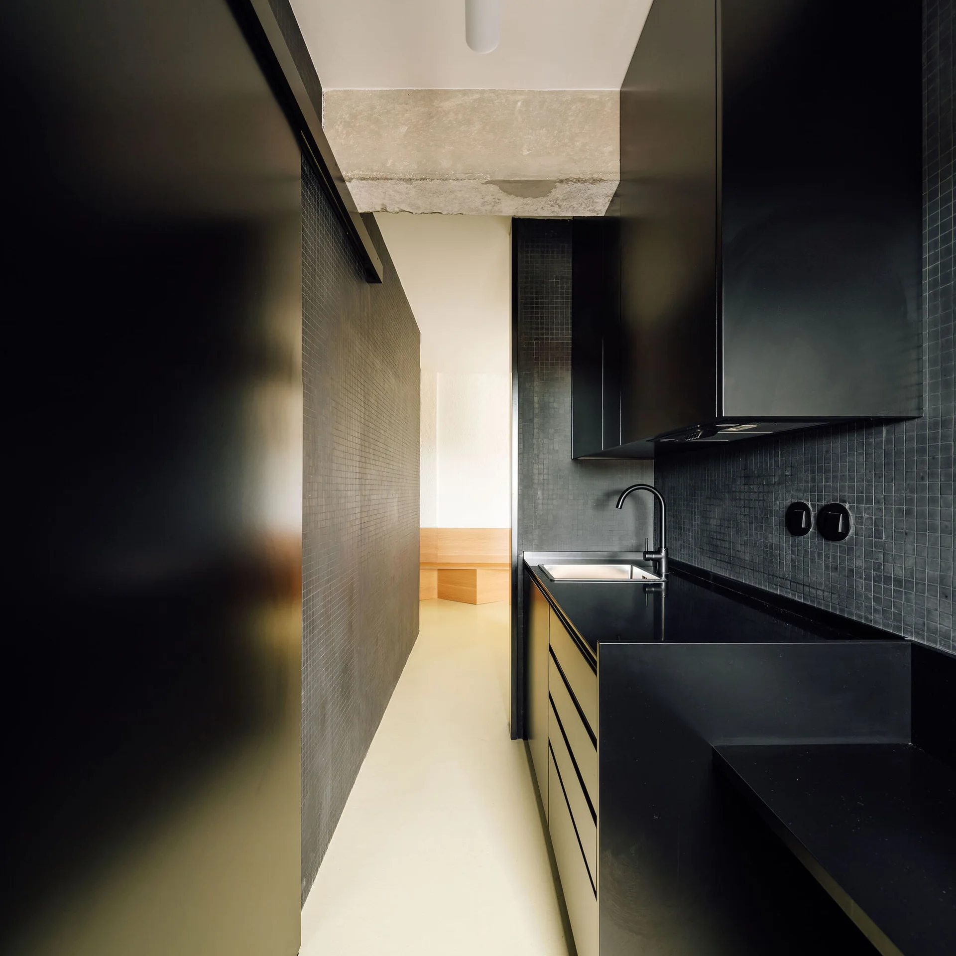 A compact modern galley kitchen featuring matte black cabinetry, black square mosaic tiled walls, and an exposed concrete ceiling beam.