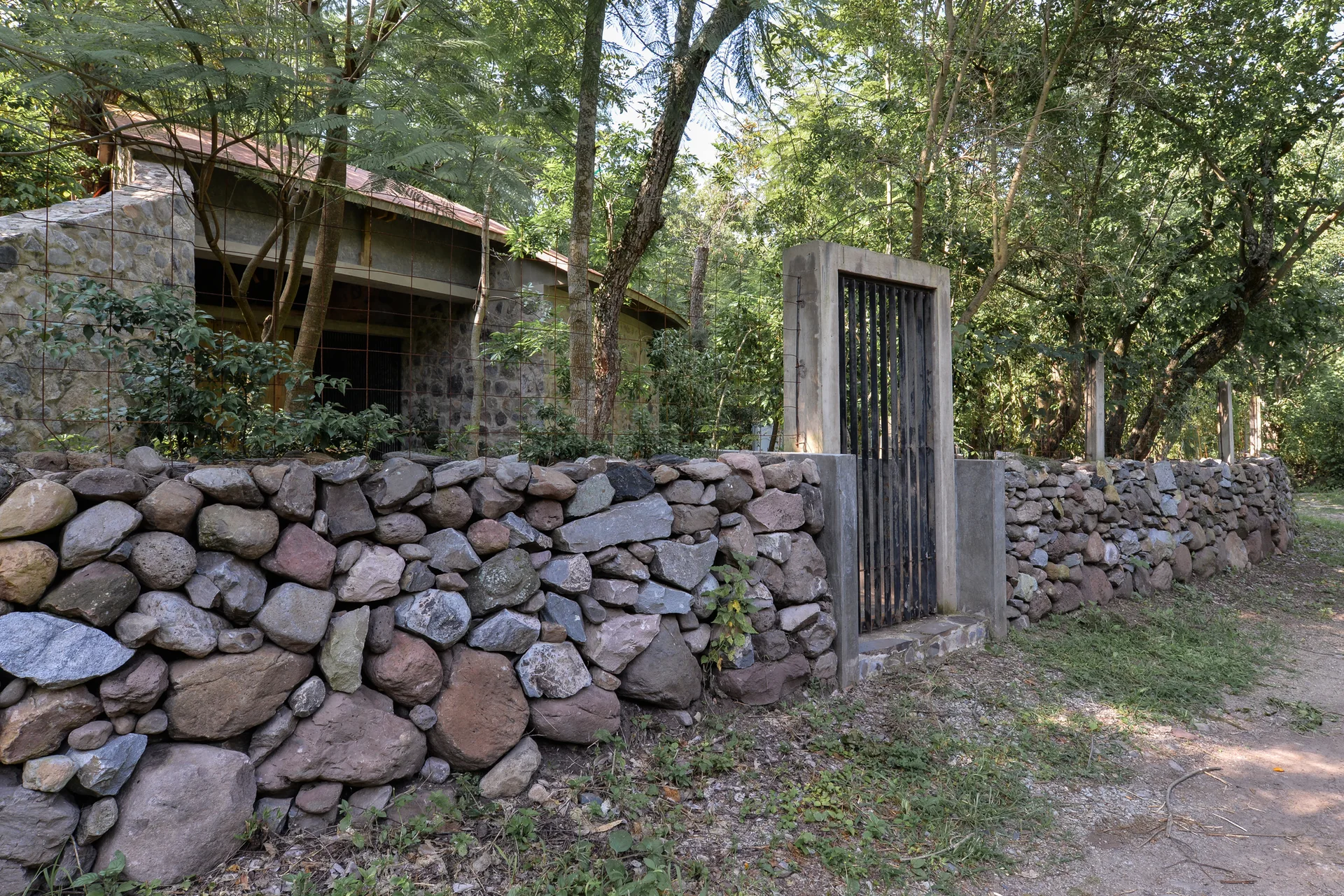 A rustic dry-stack stone wall features a black metal gate set within a concrete frame, bordered by dense green trees.