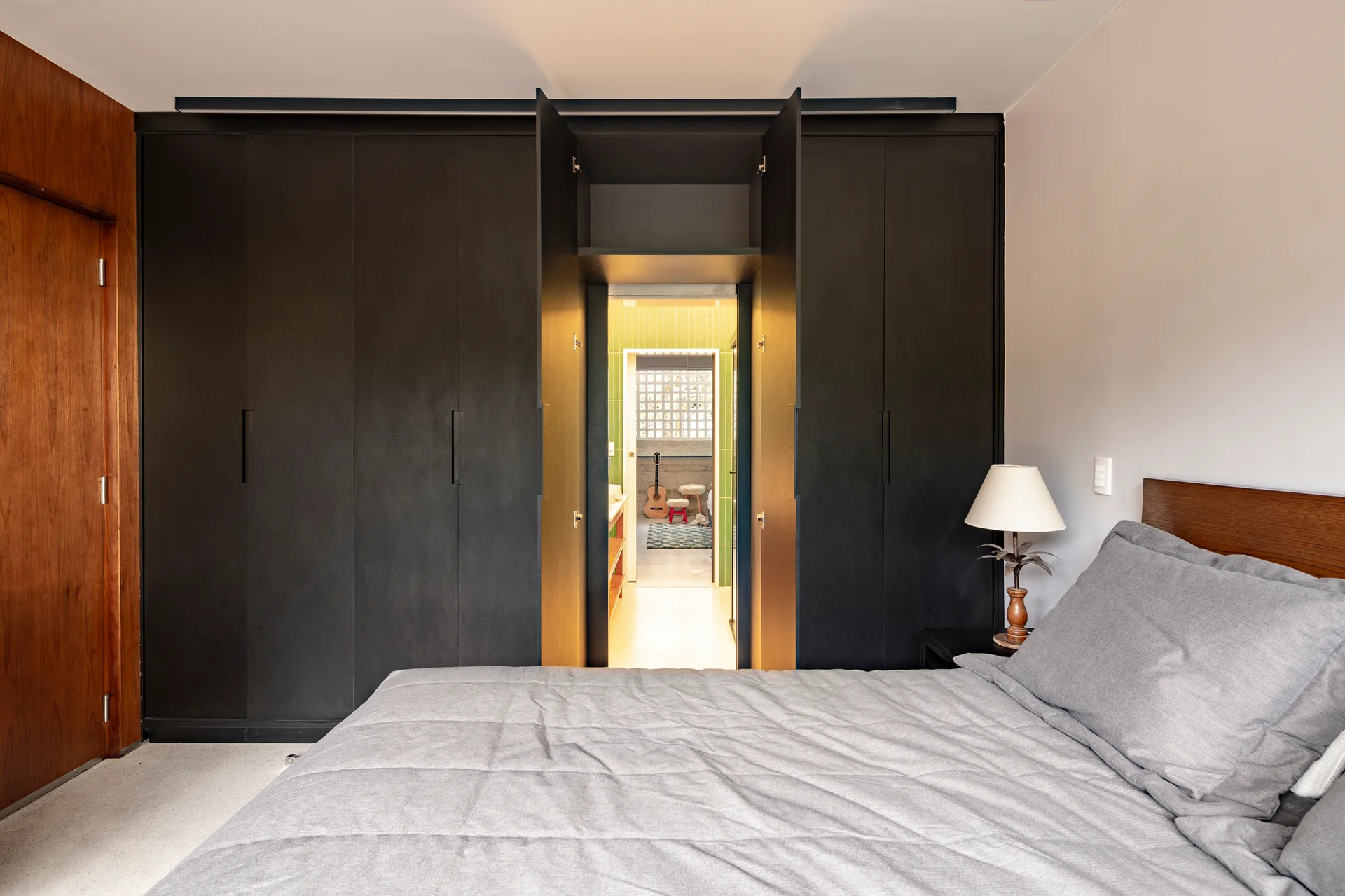 A bedroom features a grey bed next to a large matte black built-in wardrobe with a central open doorway leading to a brightly lit bathroom.