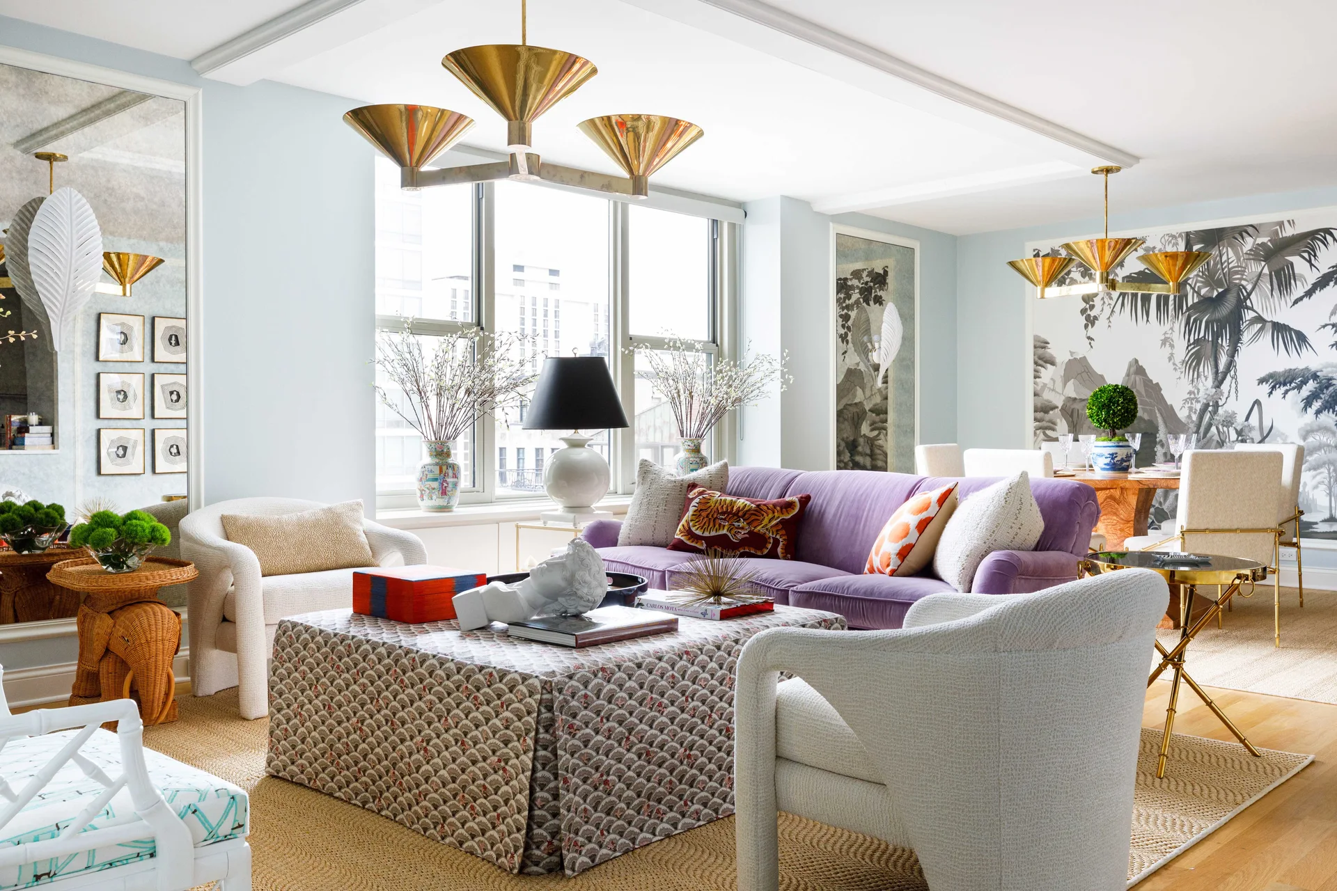 A light blue living room features a purple velvet sofa, a patterned skirted ottoman, and two white armchairs on a natural fiber rug.