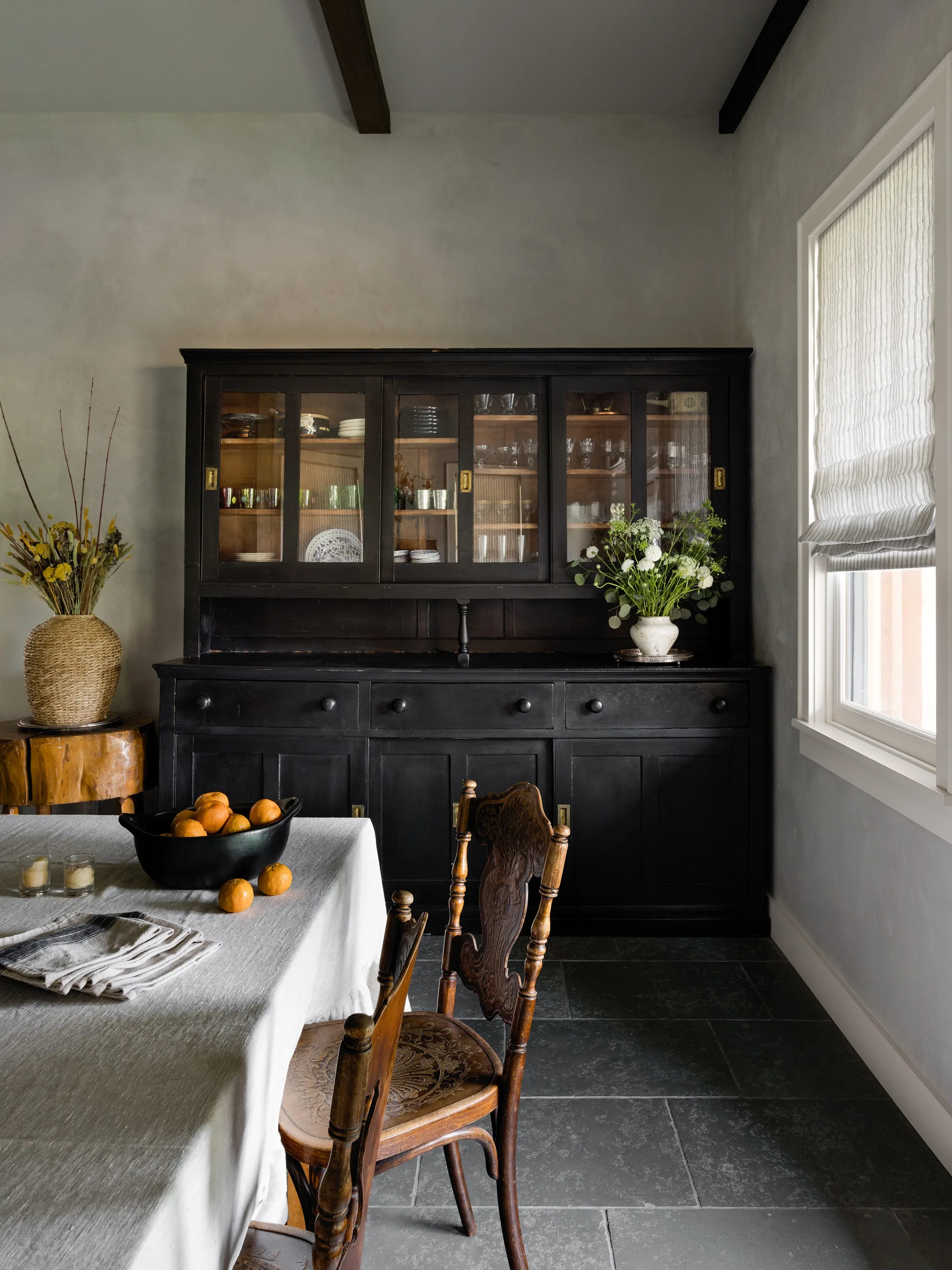 A traditional dining room featuring a large matte black hutch, a linen-covered table with a bowl of oranges, and a carved wooden chair on slate tile floors.