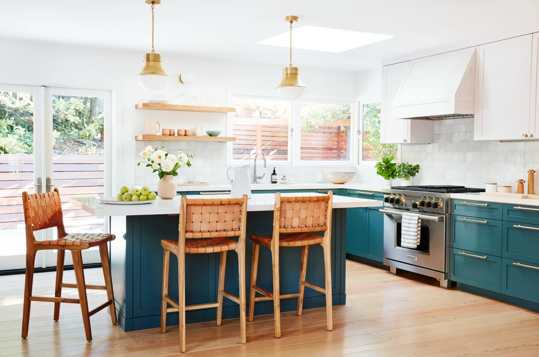A kitchen features teal lower cabinetry, a white countertop island with three woven leather stools, and two brass pendant lights hanging from the ceiling.