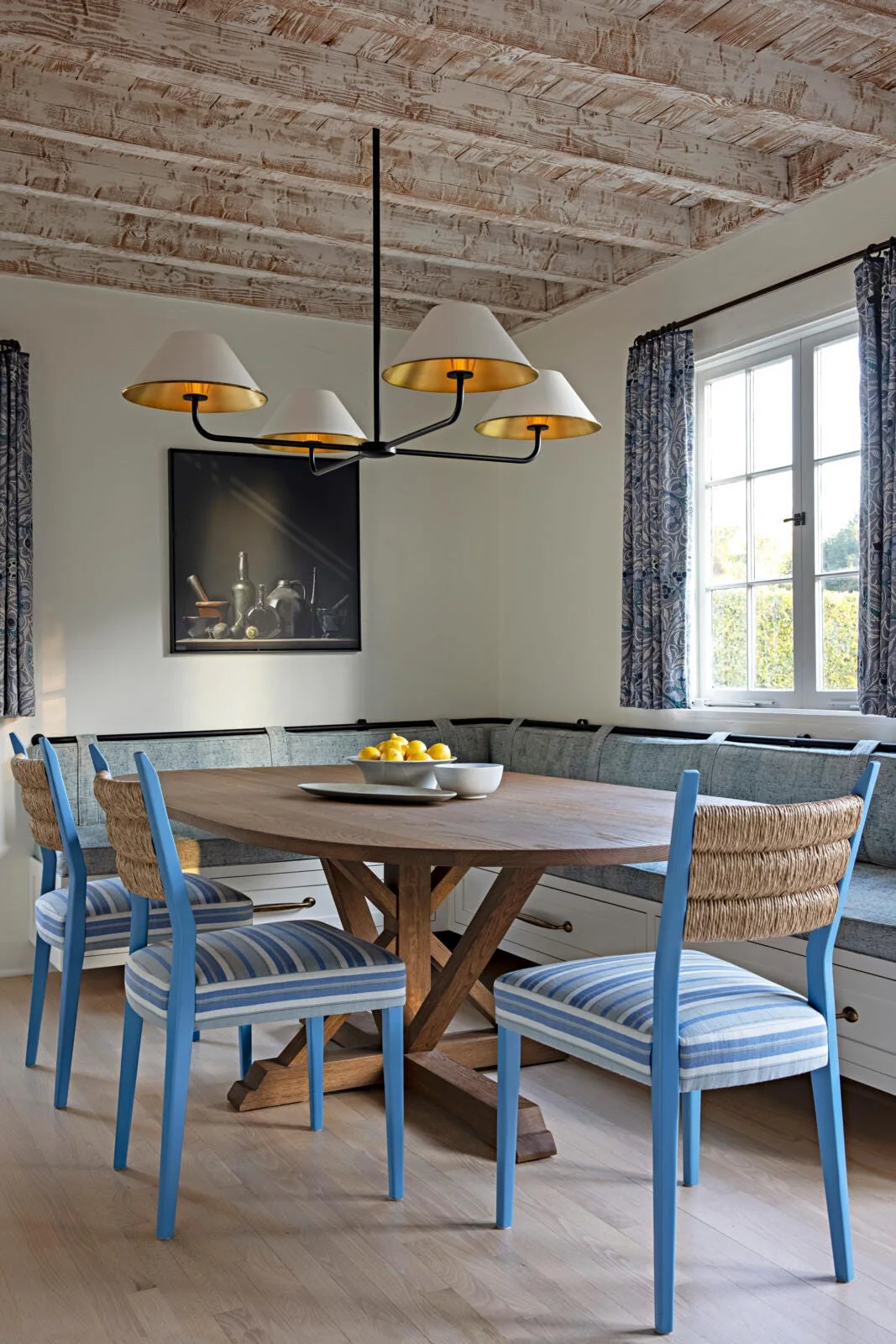A dining nook featuring a wooden table, blue painted chairs with striped cushions, a corner banquette, and a whitewashed wood beam ceiling.