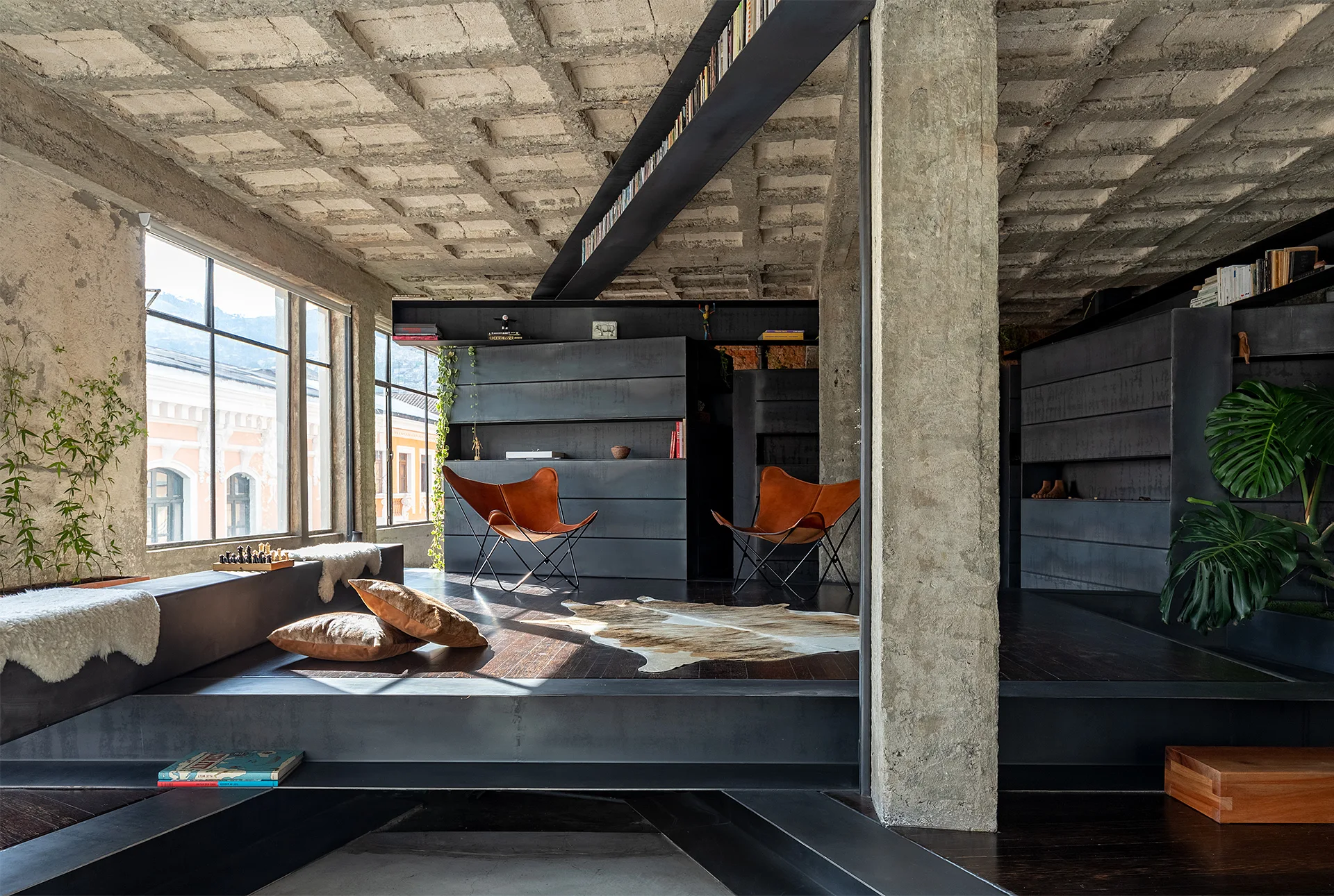 An industrial living room features an exposed concrete waffle ceiling, a structural column, dark steel built-in shelving, and two tan leather butterfly chairs.