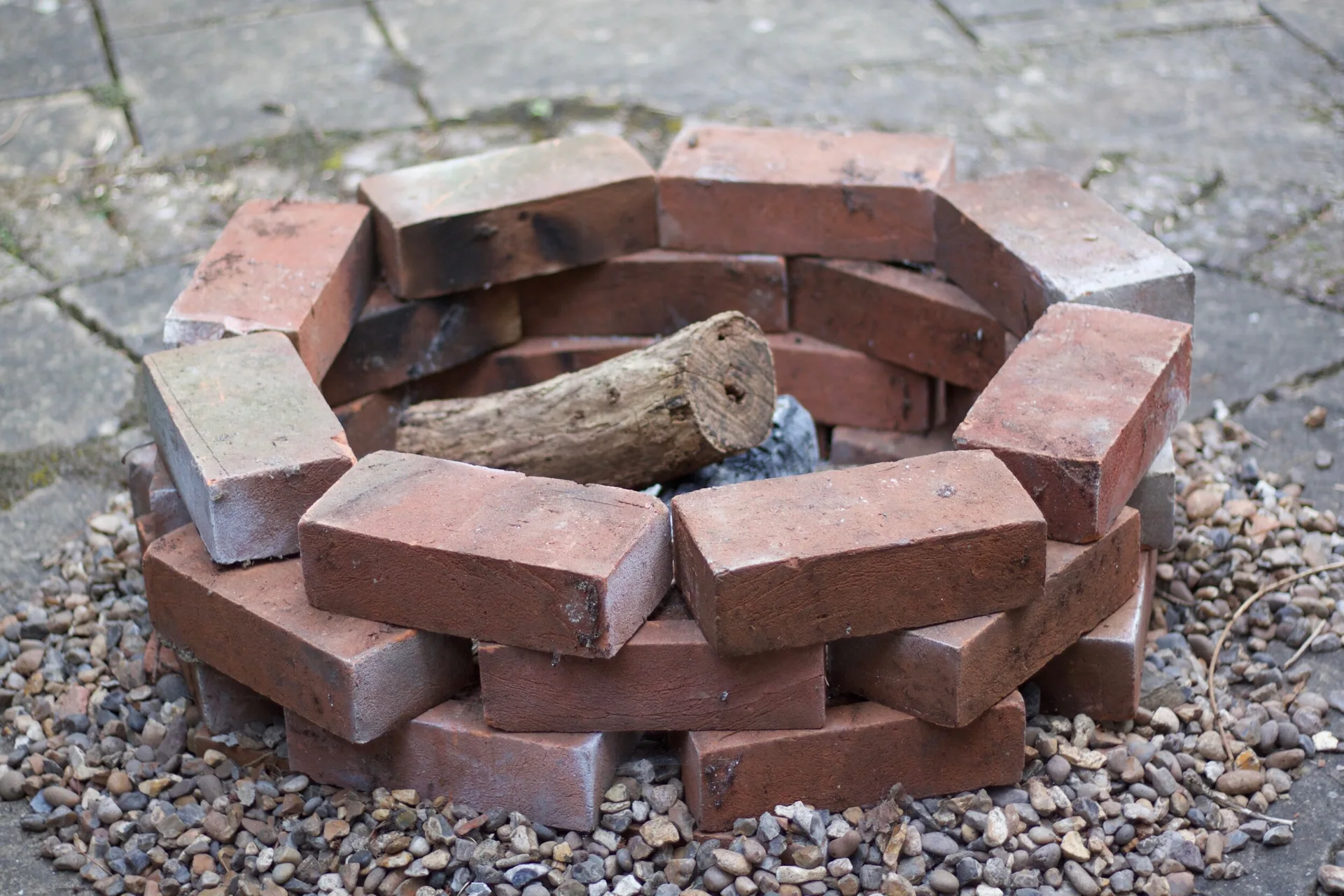 A small circular fire pit constructed from stacked red bricks sits on a stone patio surrounded by grey gravel with a single log inside.