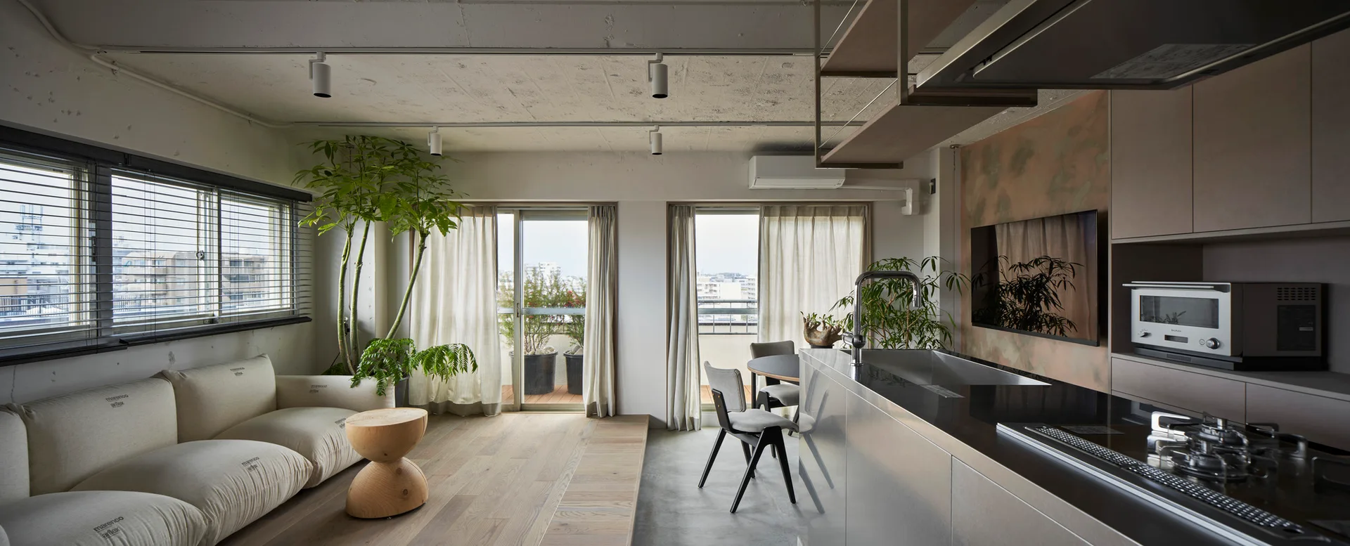 A modern open-plan living and kitchen space featuring a raised wooden platform floor, concrete ceiling, and modular cream sofa.