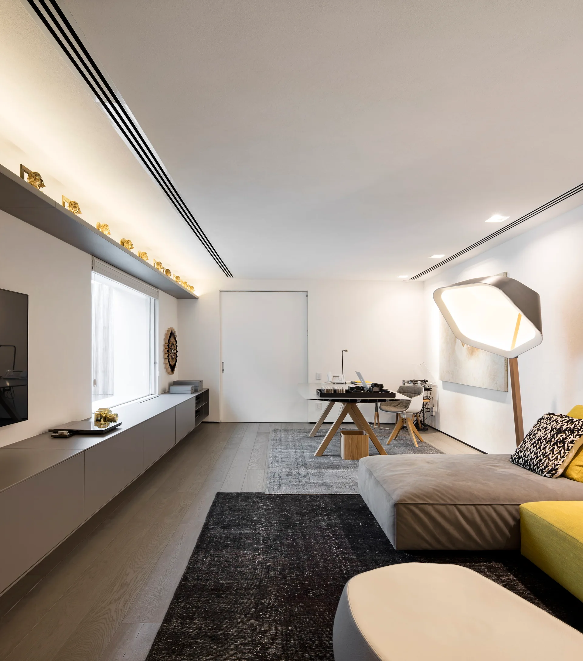 A modern living room features a modular sofa with yellow accents, low grey cabinetry along the wall, and a desk area under recessed ceiling lights.