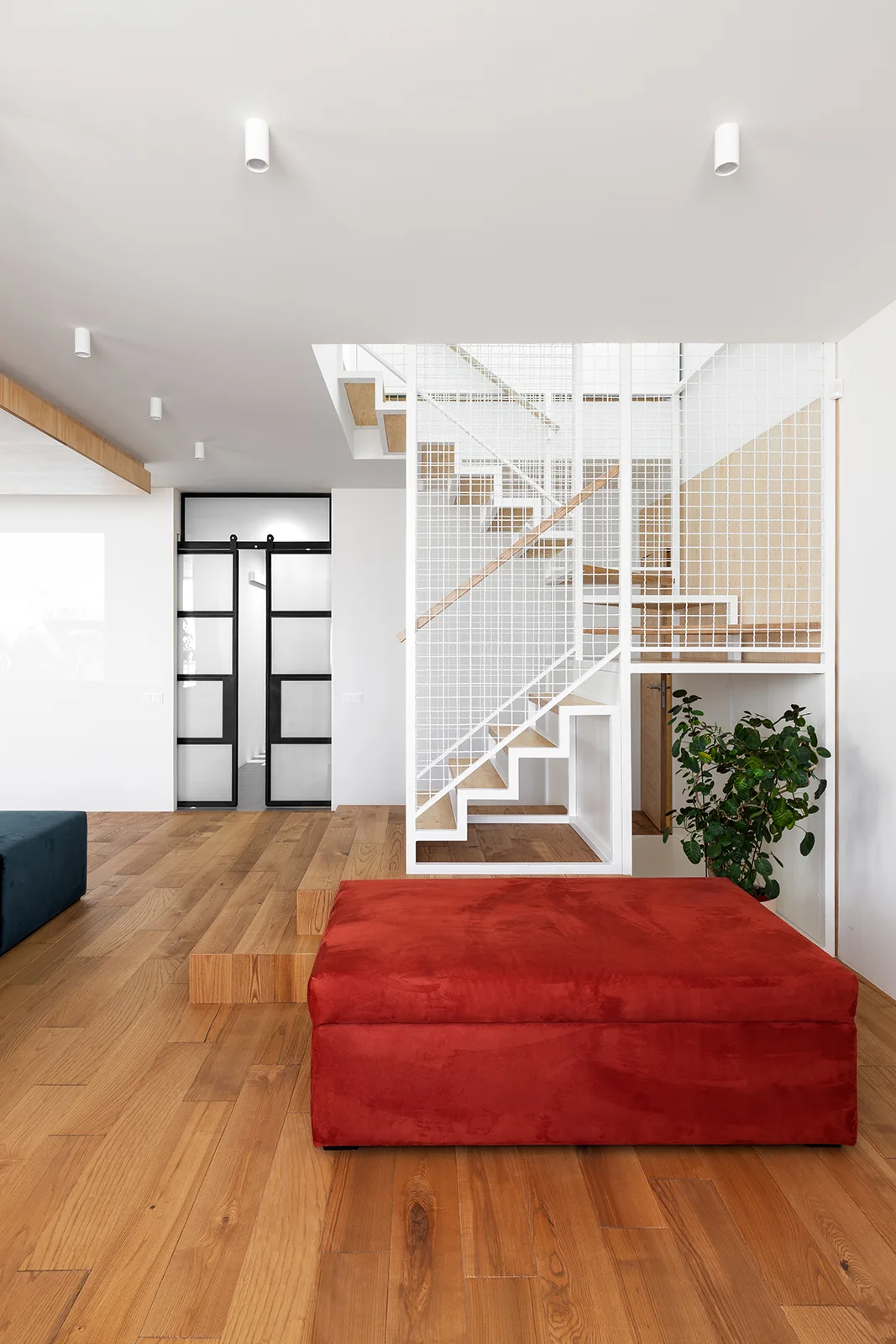 A modern home interior features wood floors, a large red ottoman, and a staircase with a white metal grid safety railing.