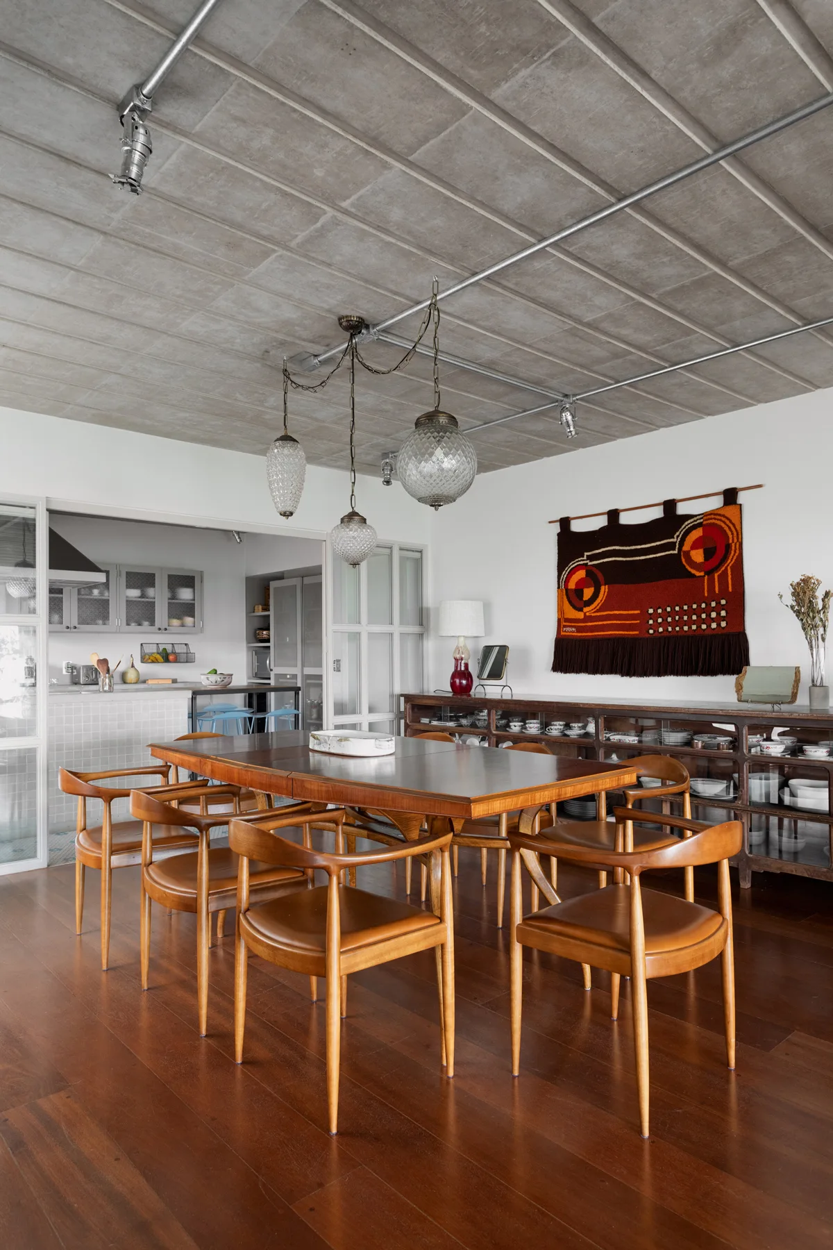 A mid-century modern wooden dining set under an exposed concrete ceiling with glass pendant lights and a woven wall tapestry.