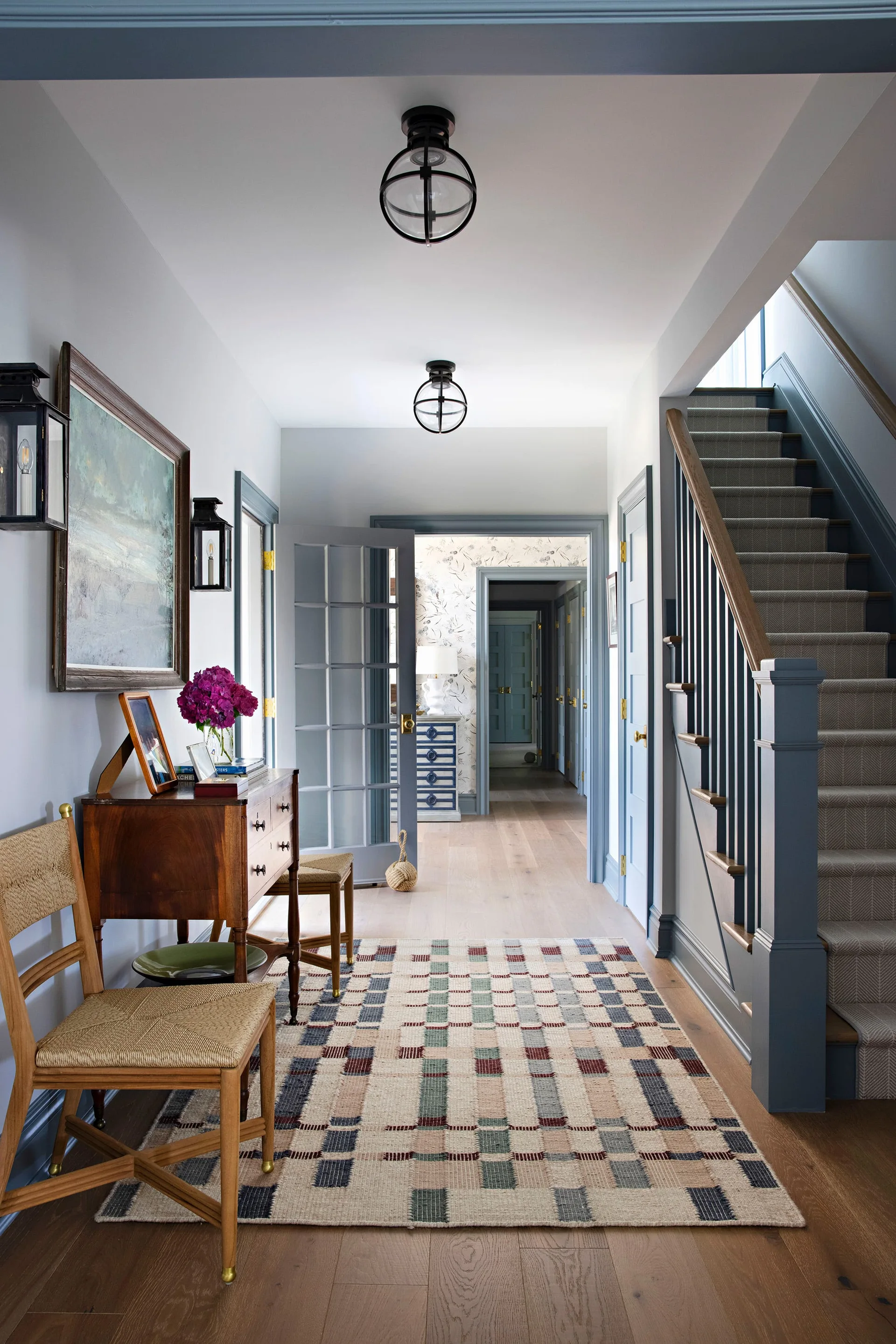 A traditional foyer featuring slate blue painted trim, an antique wood console table with woven chairs, and a staircase with a striped runner.