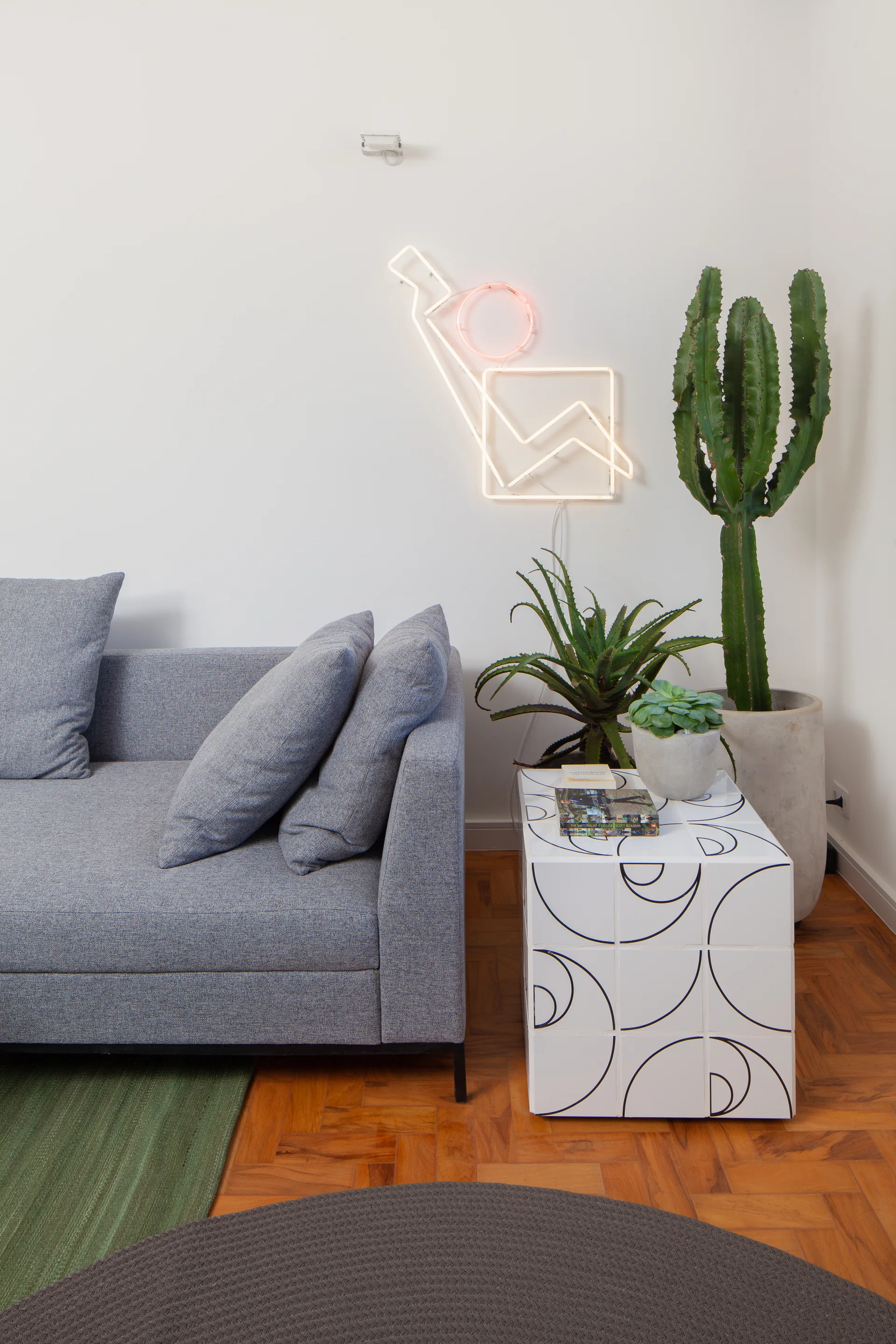 A grey fabric sofa sits beside a tiled cube side table with black geometric patterns, a tall potted cactus, and an abstract neon wall light.