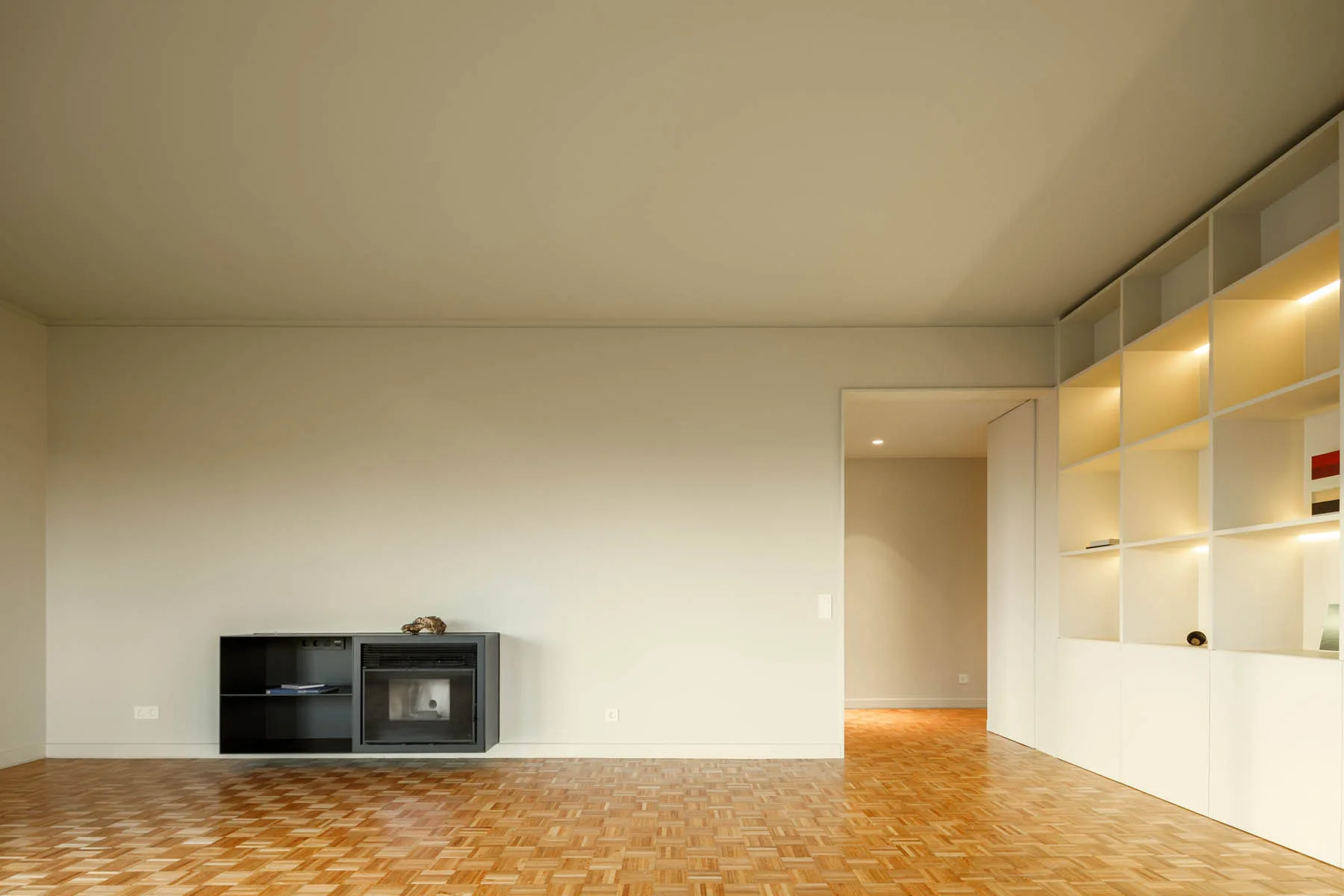 A minimalist room featuring wood parquet flooring, a low-profile black fireplace console, and built-in white shelving with warm integrated lighting.