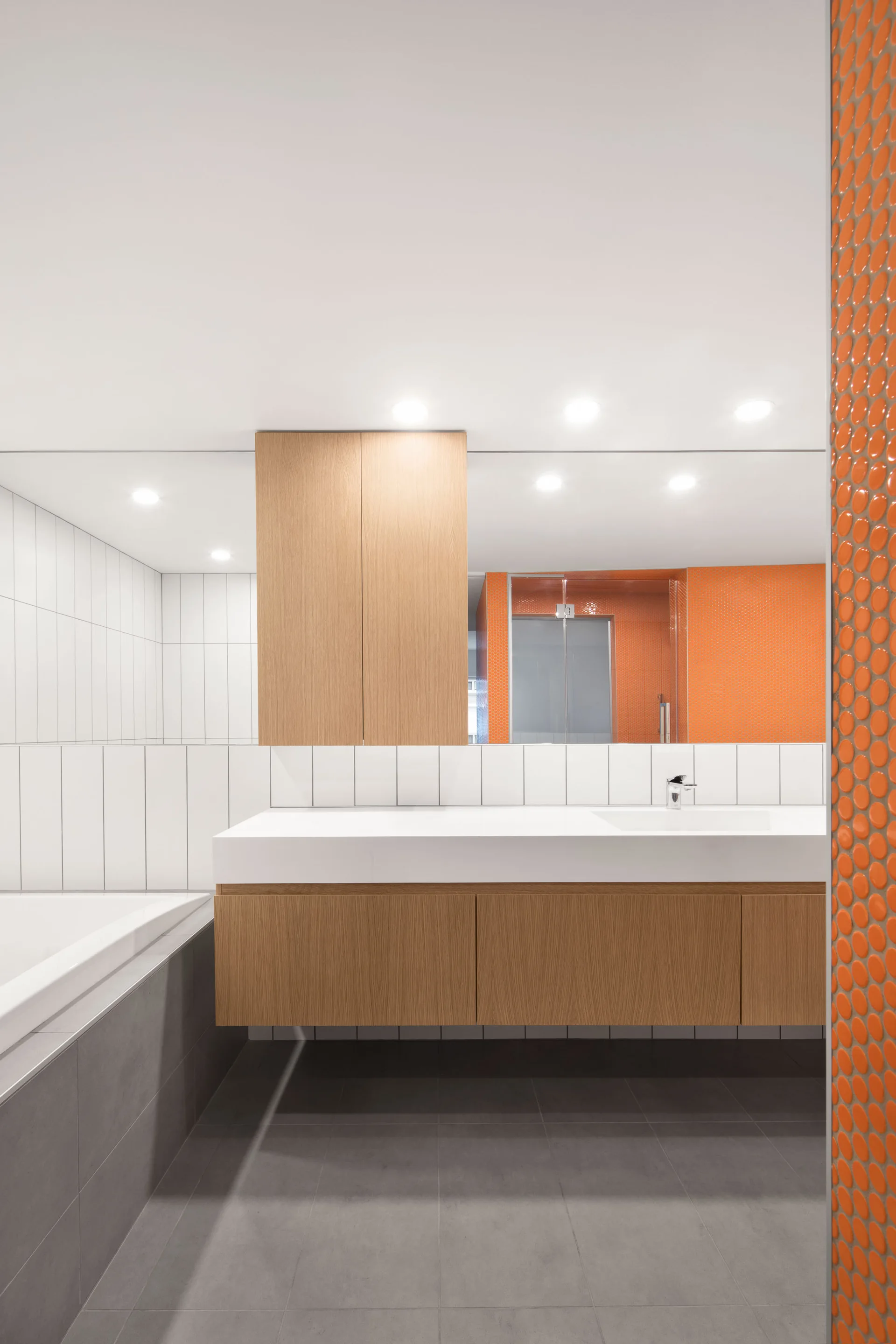 A modern bathroom features a wood-toned floating vanity, a large wall mirror, white vertical tiles, grey floor tiles, and a bright orange mosaic tile accent.