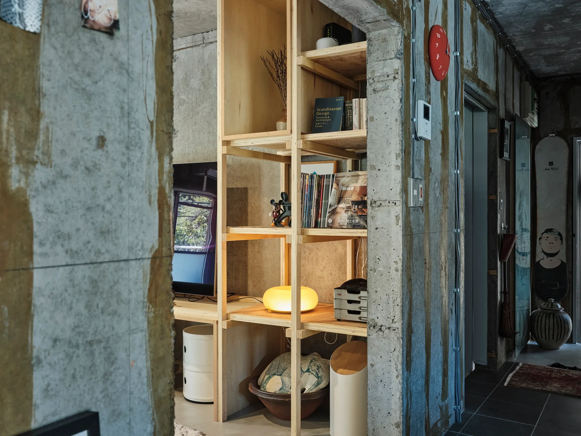 An open plywood shelving unit stands against exposed concrete walls in an industrial-style room featuring a glowing orb lamp and books.