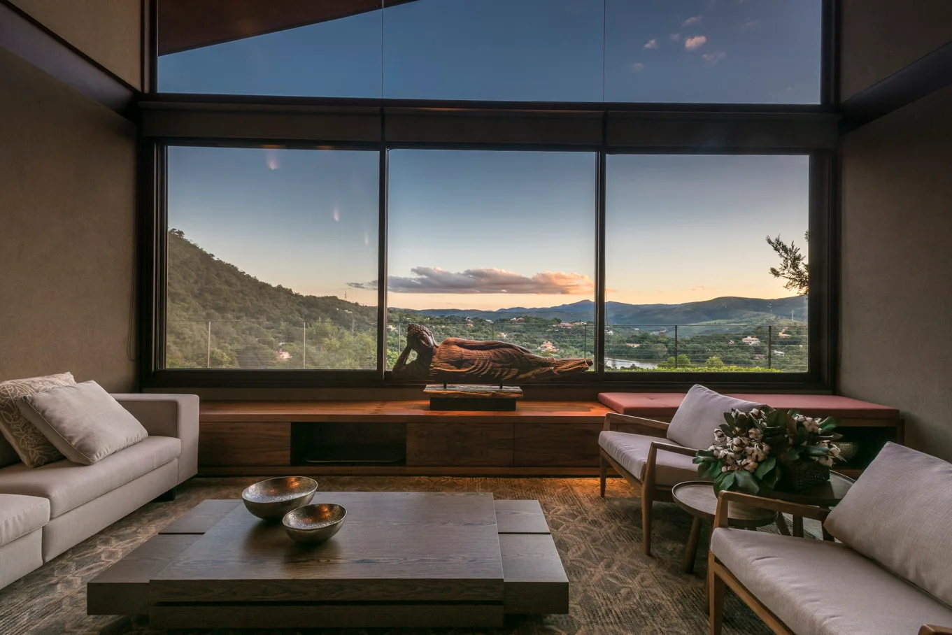A living room with low wooden furniture features a large panoramic window overlooking mountain views at sunset behind a reclining Buddha sculpture.