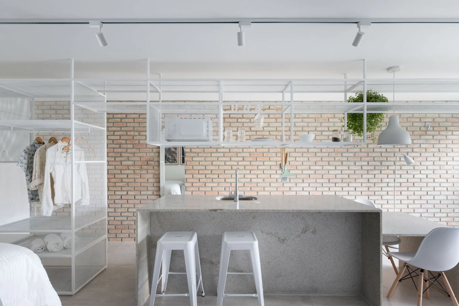 A modern kitchen area featuring a gray stone island with white stools, a white metal mesh storage rack, and an exposed brick wall background.