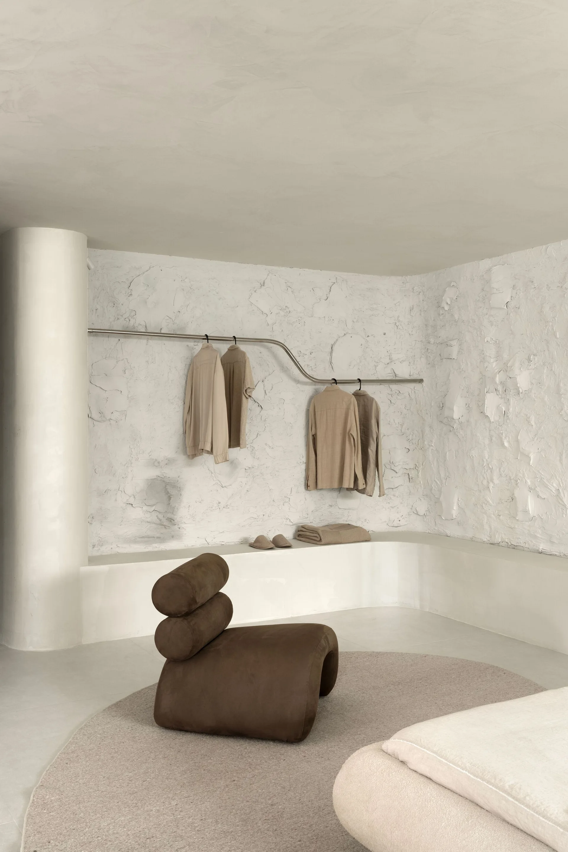A minimalist dressing space with rough white plaster walls, a curved metal clothing rail, a brown sculptural chair, and a low plaster ledge.