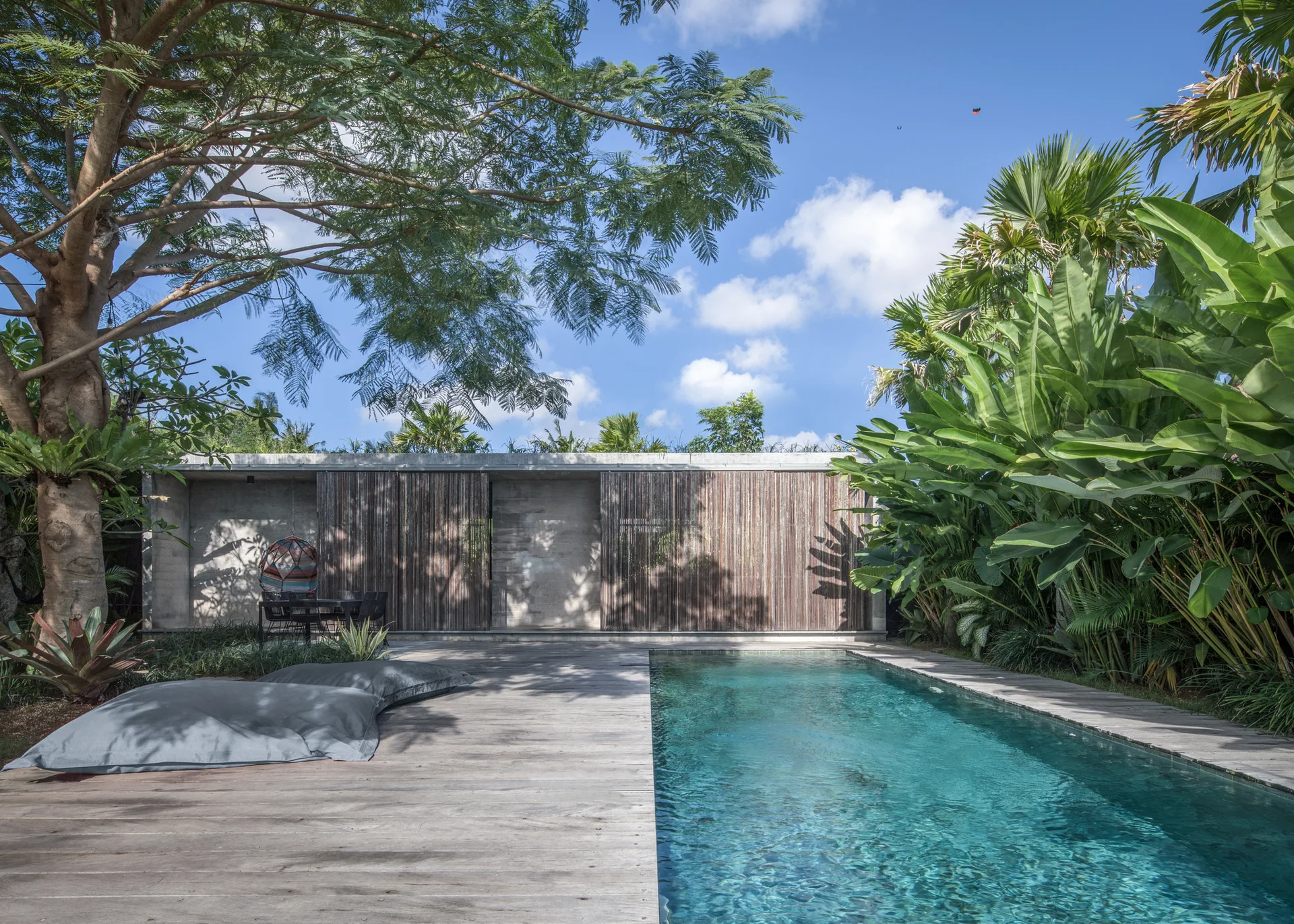 A rectangular turquoise swimming pool is bordered by weathered wood decking and a raw concrete building with sliding slatted wooden screens.