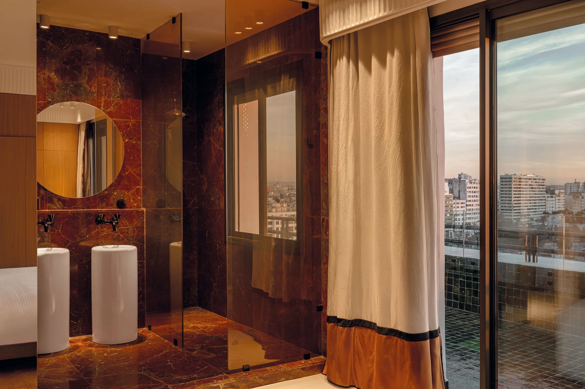 A modern bathroom featuring dark brown veined marble walls, two cylindrical white pedestal sinks, a round mirror, and a large sliding glass balcony door.