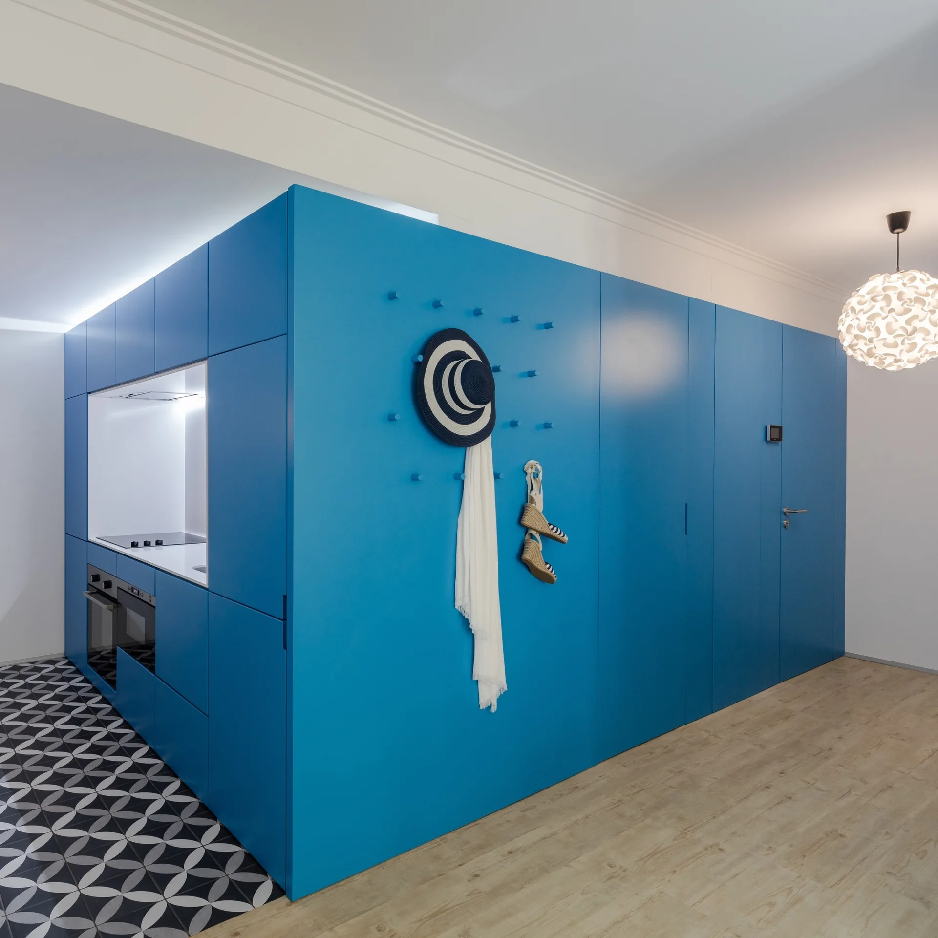 A large blue cabinet unit houses a recessed kitchen niche over patterned geometric floor tiles next to a light wood floor.
