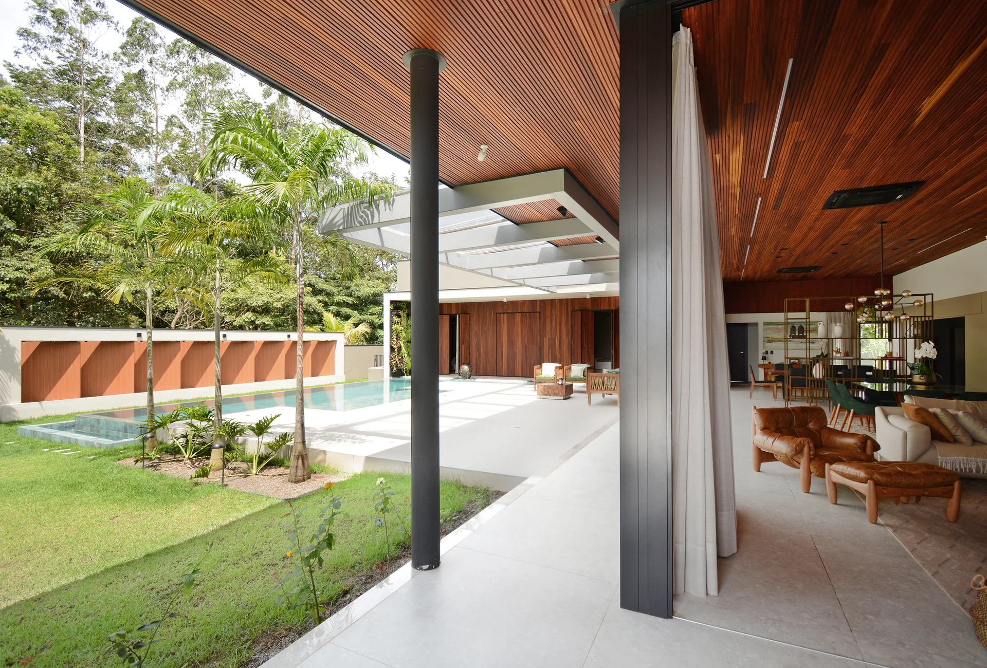 A modern open-plan living room with a slatted wood ceiling transitions seamlessly onto a concrete patio with a swimming pool and lawn.