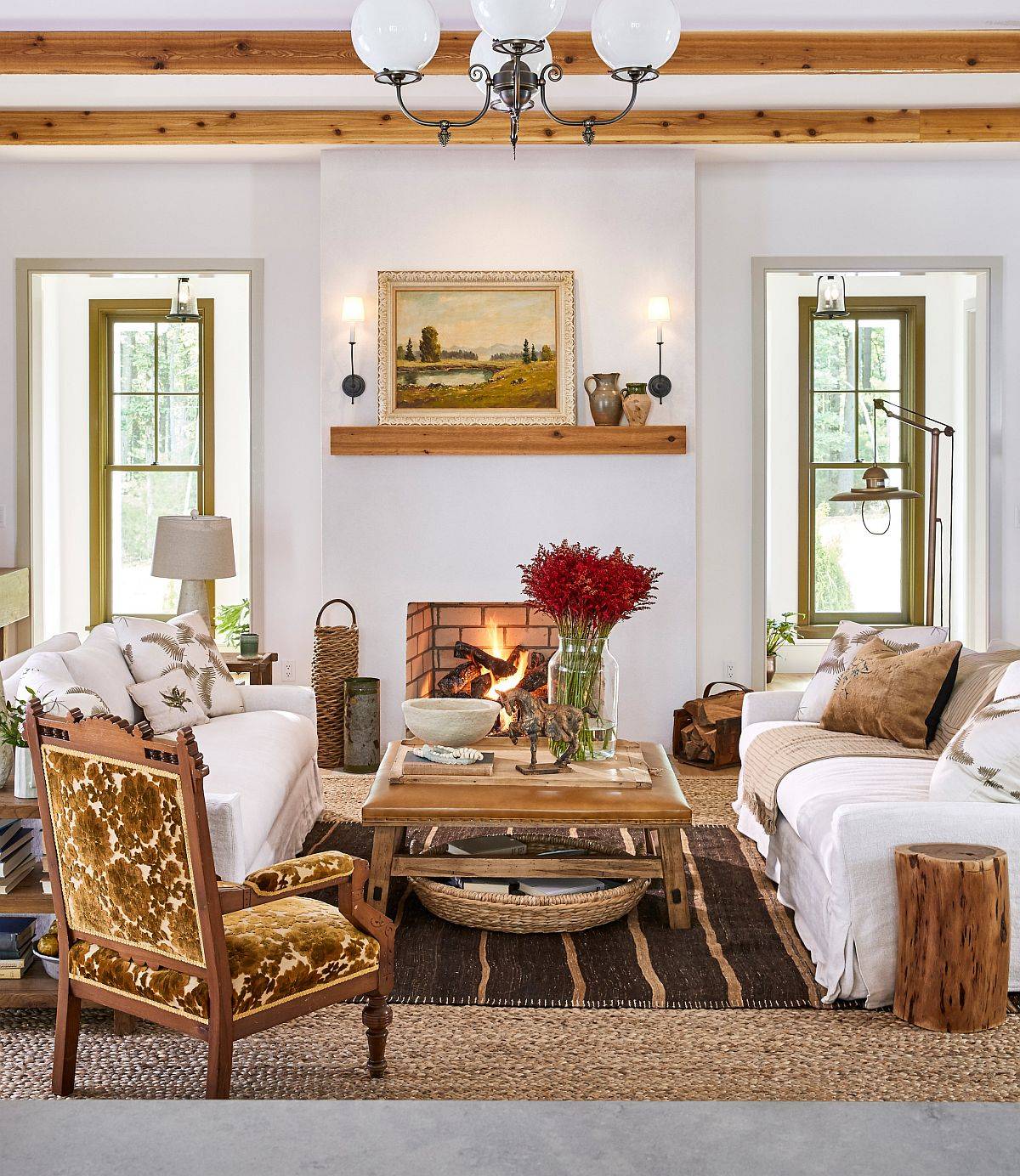 A cozy rustic living room featuring a brick fireplace, white slipcovered sofas, olive green window frames, and exposed wood ceiling beams.