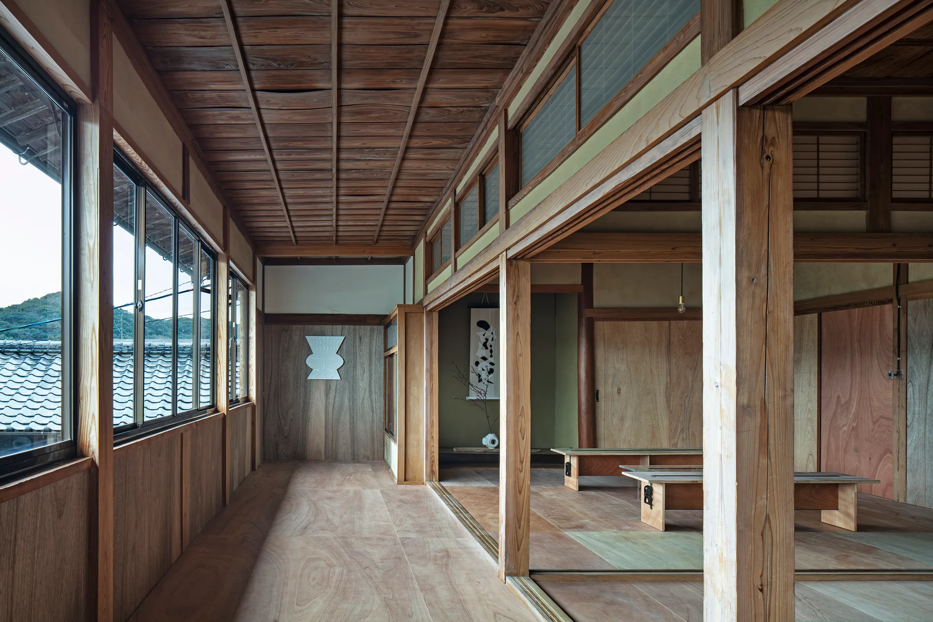 A traditional Japanese corridor features natural wood paneling, sliding partitions, low wooden benches, and large windows overlooking tiled roofs.
