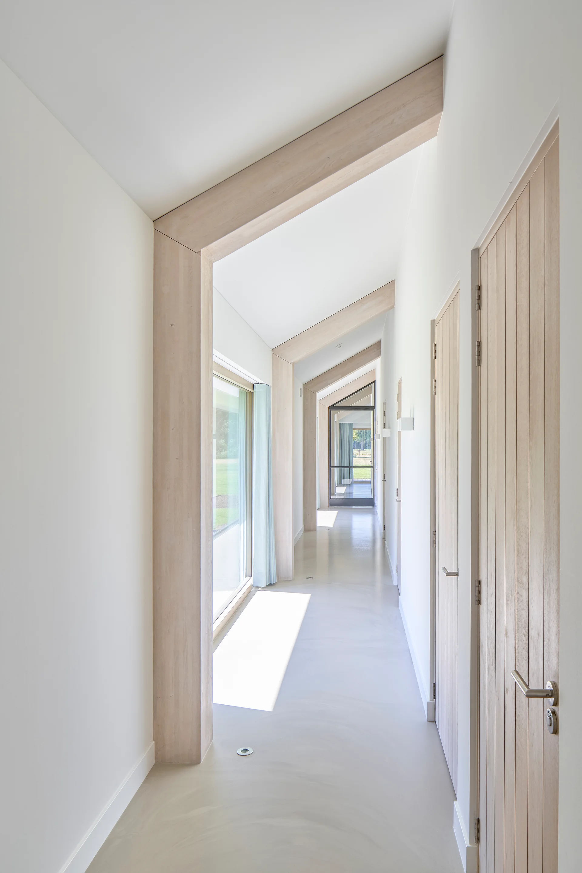 A long minimalist hallway with exposed light wood structural beams, vertical wood-paneled doors, white walls, and smooth light-colored floors.