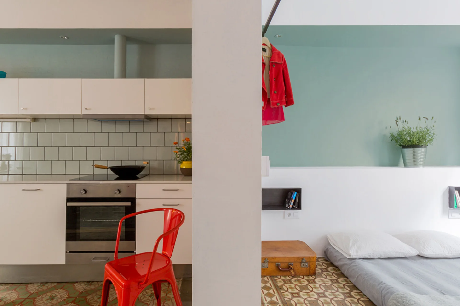 A studio apartment interior divided by a white pillar features a white kitchen on the left and a floor bed on the right, set on patterned tile floors.