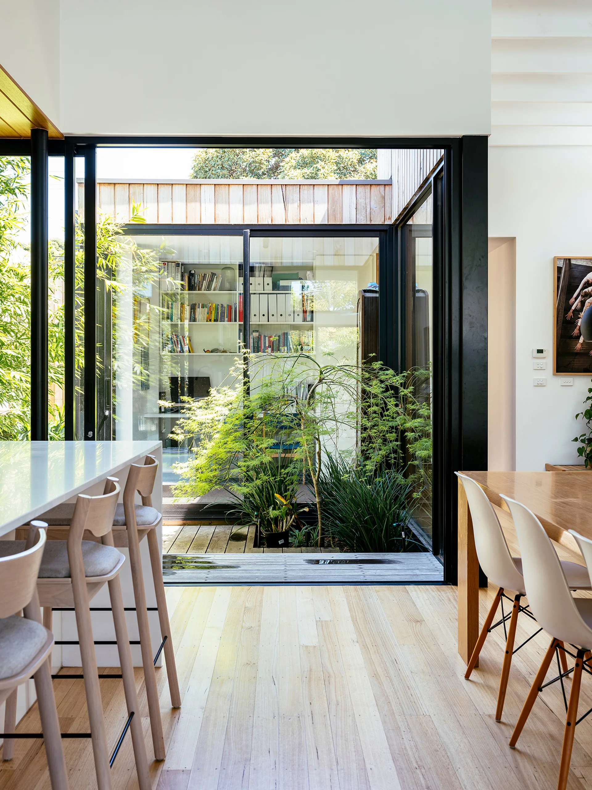 A light wood dining table and kitchen island flank large black-framed sliding glass doors that open to a small green courtyard garden.