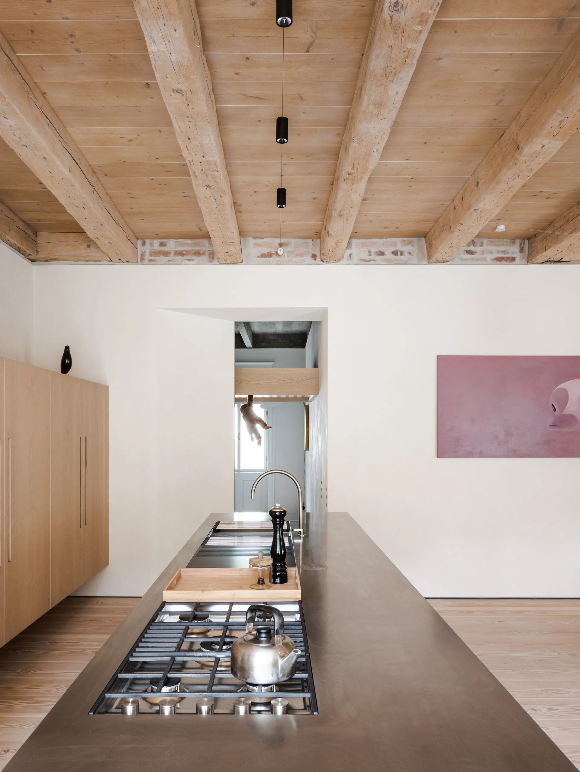 A minimalist kitchen features a long gray island with a gas cooktop, set beneath exposed wooden ceiling beams and light wood cabinets.