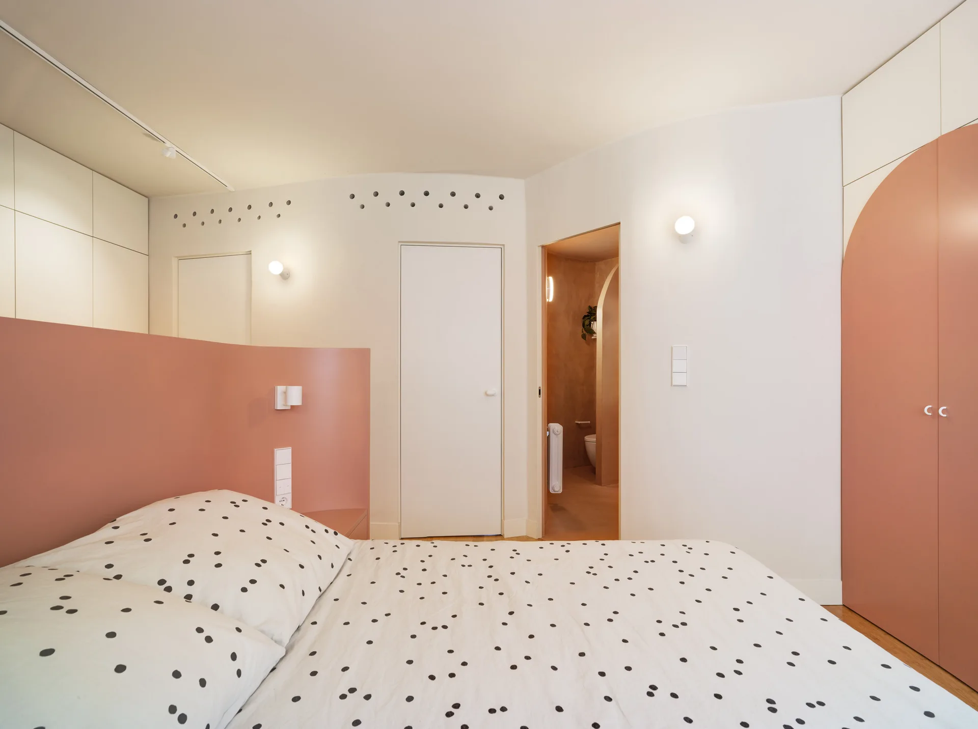 A modern bedroom with polka dot bedding features a curved dusty pink headboard panel, white walls, and a built-in pink wardrobe with arched detailing.