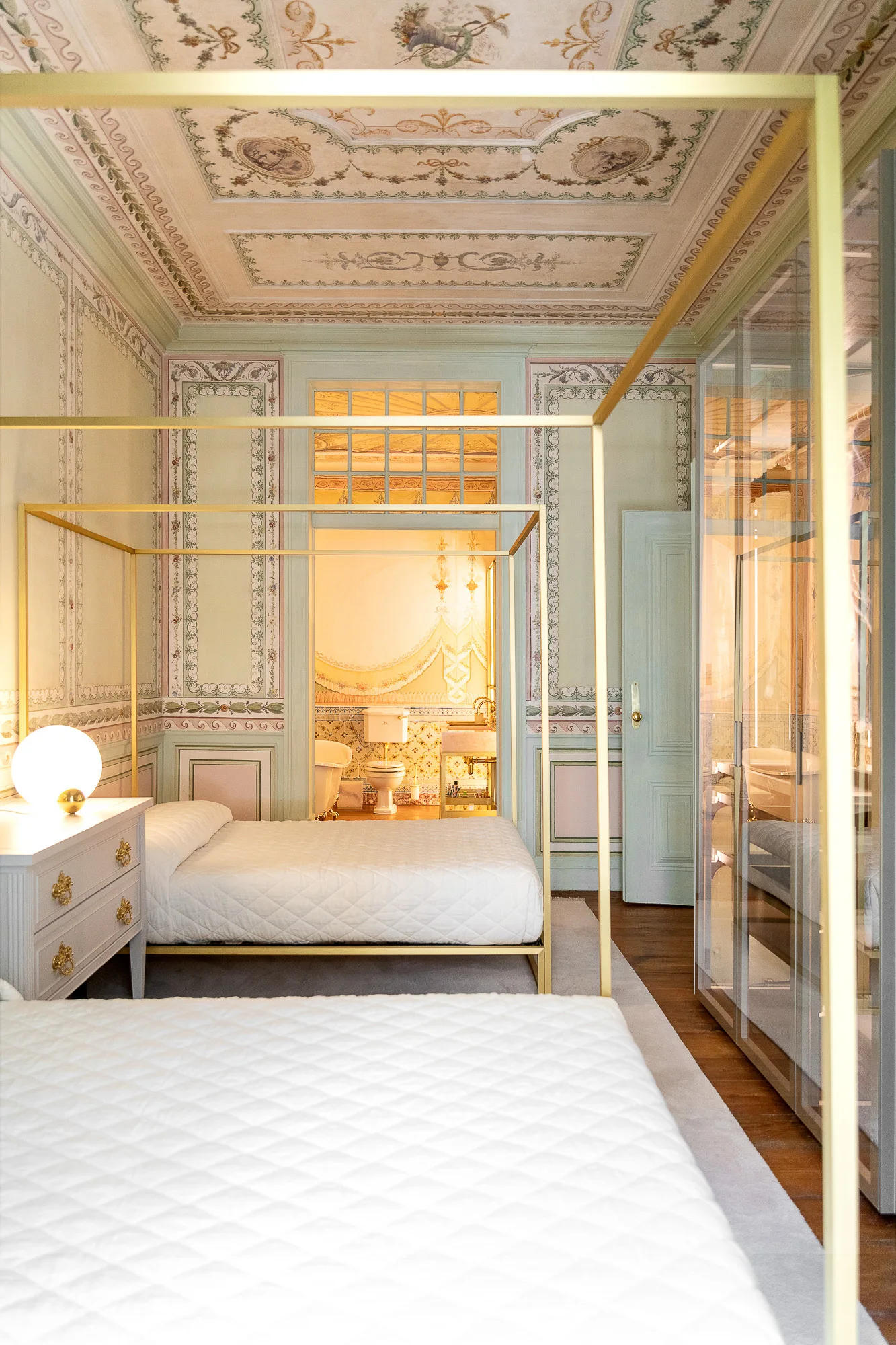 A pale green bedroom features two gold metal canopy beds with white bedding set against ornate painted walls and an elaborate fresco ceiling.