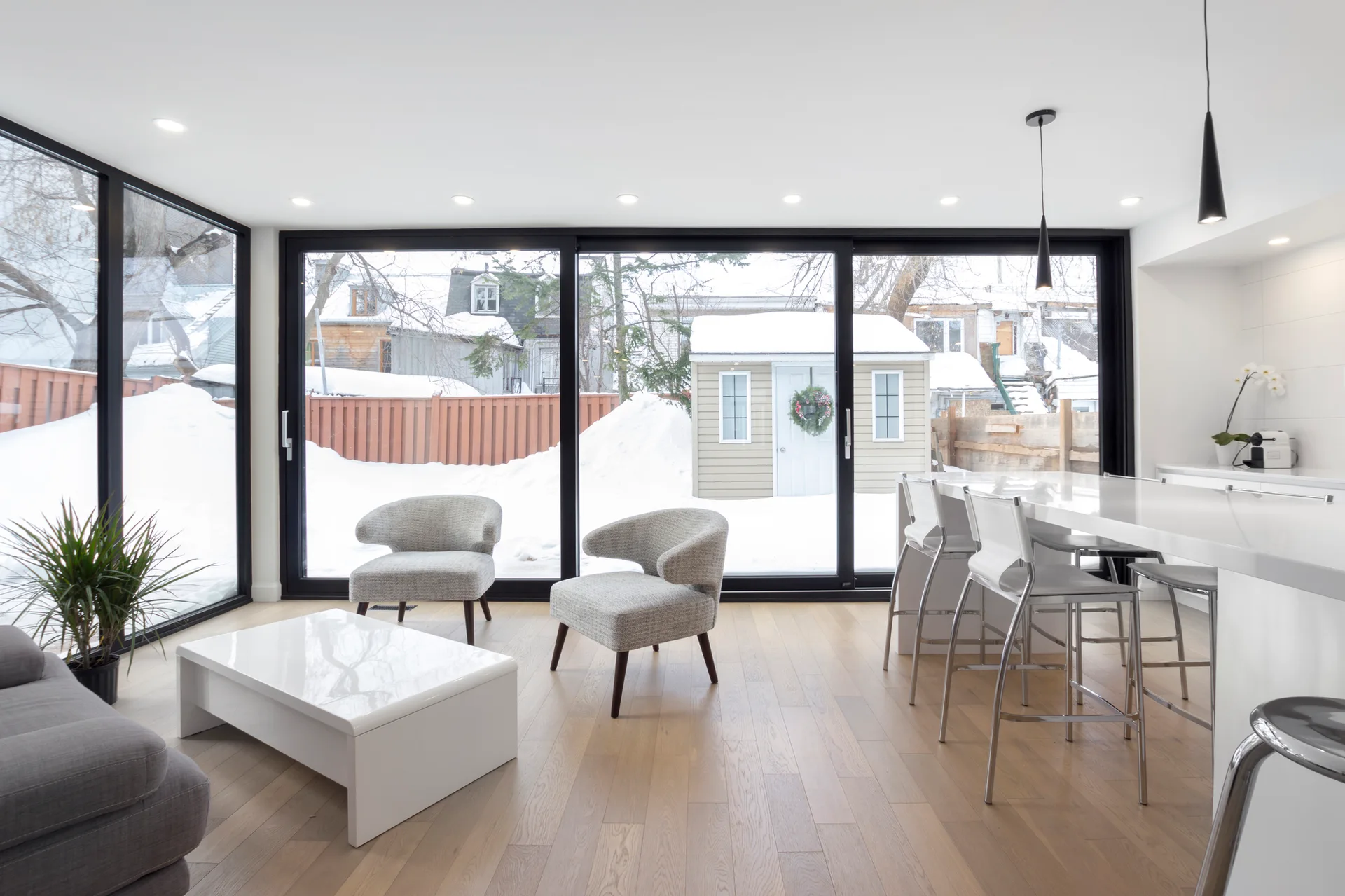 A modern open-concept living room features two gray armchairs, a white coffee table, and large black-framed sliding glass doors showing a snowy yard.