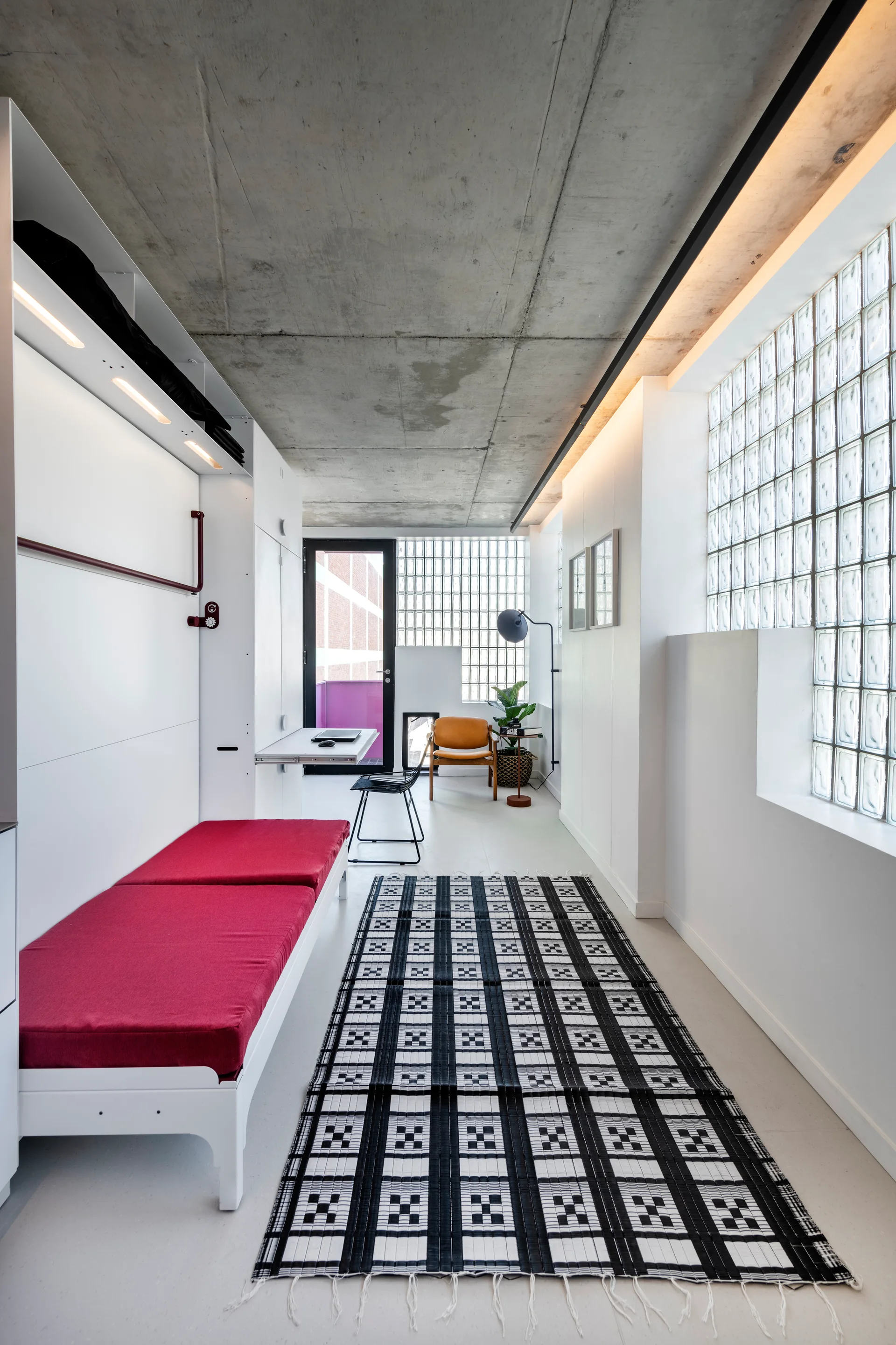 A narrow modern studio featuring an exposed concrete ceiling, a black and white geometric grid rug, and a glass block window wall.