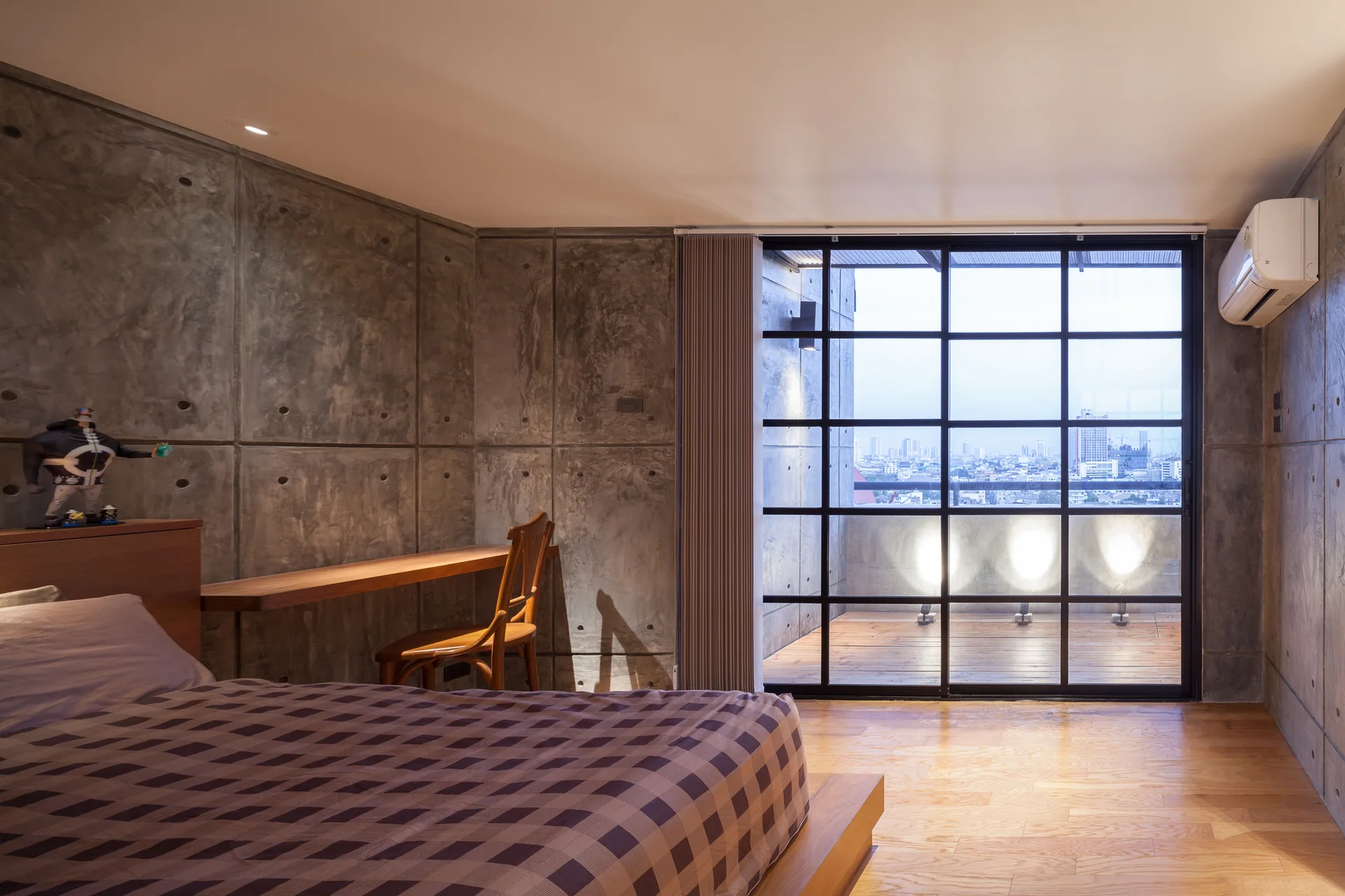 An industrial bedroom features grey concrete wall panels, a low-profile bed, a floating wooden desk with a chair, and a large black grid window.