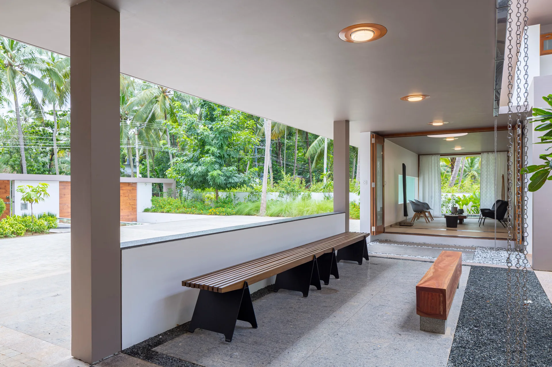 A covered modern patio features a long slatted wooden bench and a solid wood bench on stone tile flooring, looking out toward a tropical garden.