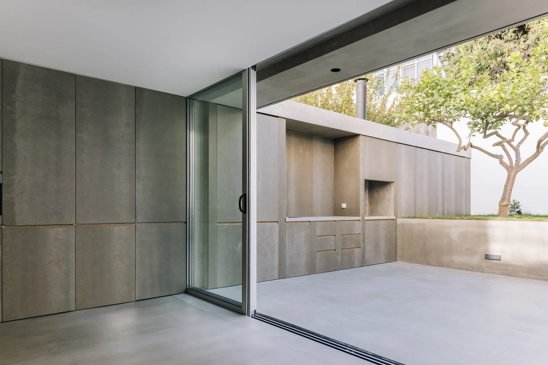 A grey concrete outdoor kitchen with built-in cabinetry sits next to a large sliding glass door on a minimalist patio with a small tree.