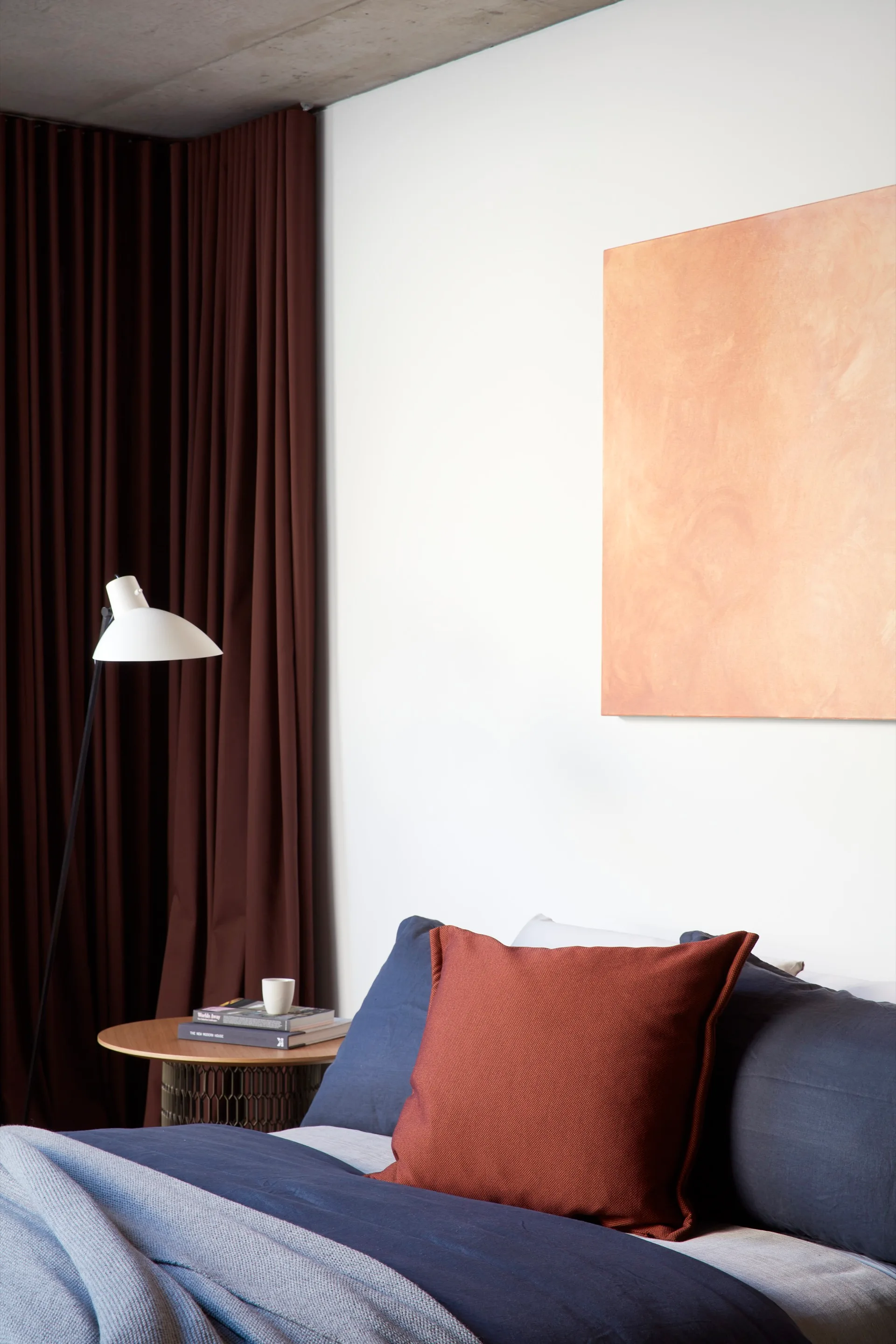A modern bedroom corner featuring blue bedding, a terracotta throw pillow, a textured orange canvas painting, and deep brown curtains.