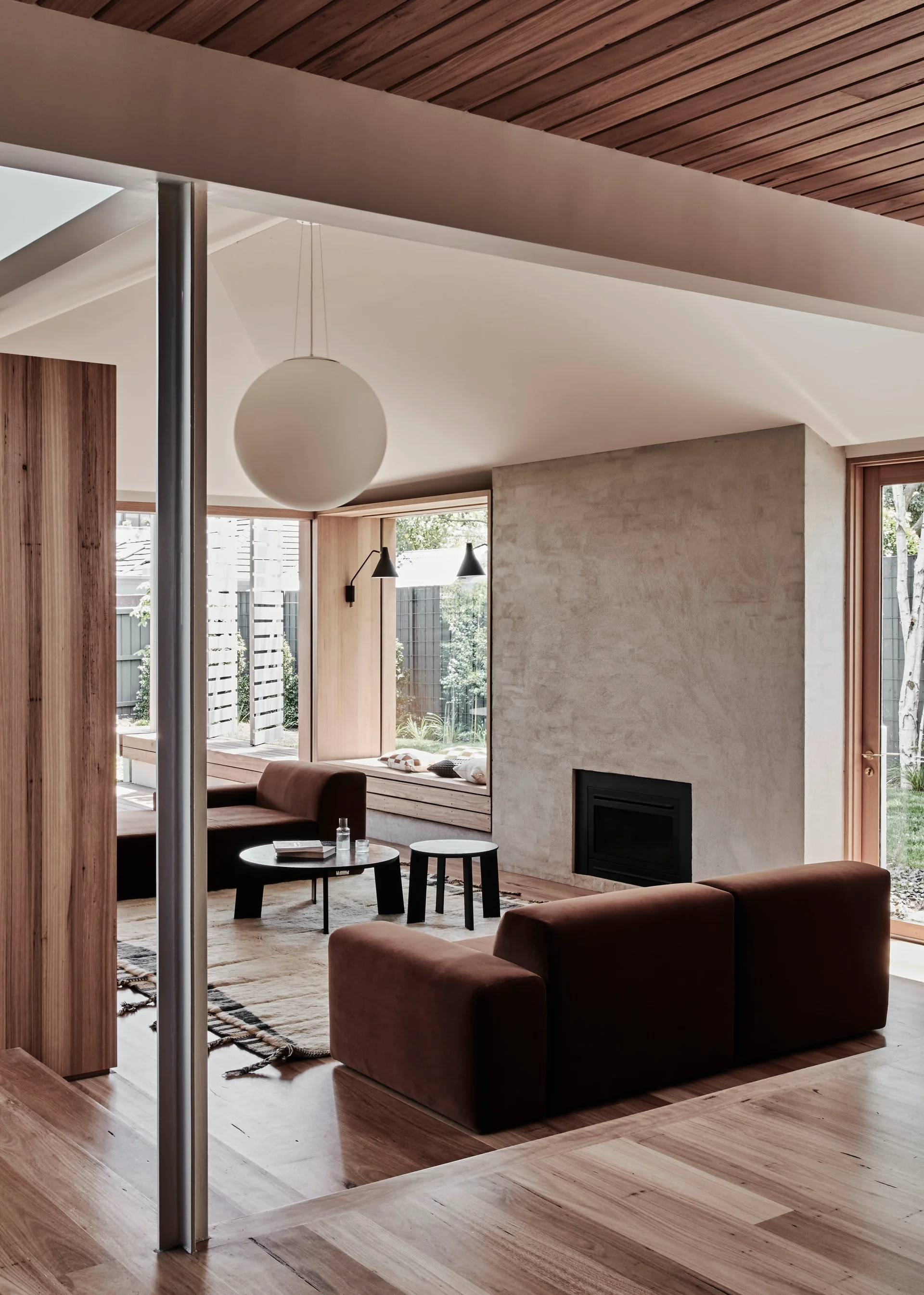 A modern living room featuring a brown modular sofa, a textured plaster fireplace, a slatted timber ceiling, and a large spherical pendant light.