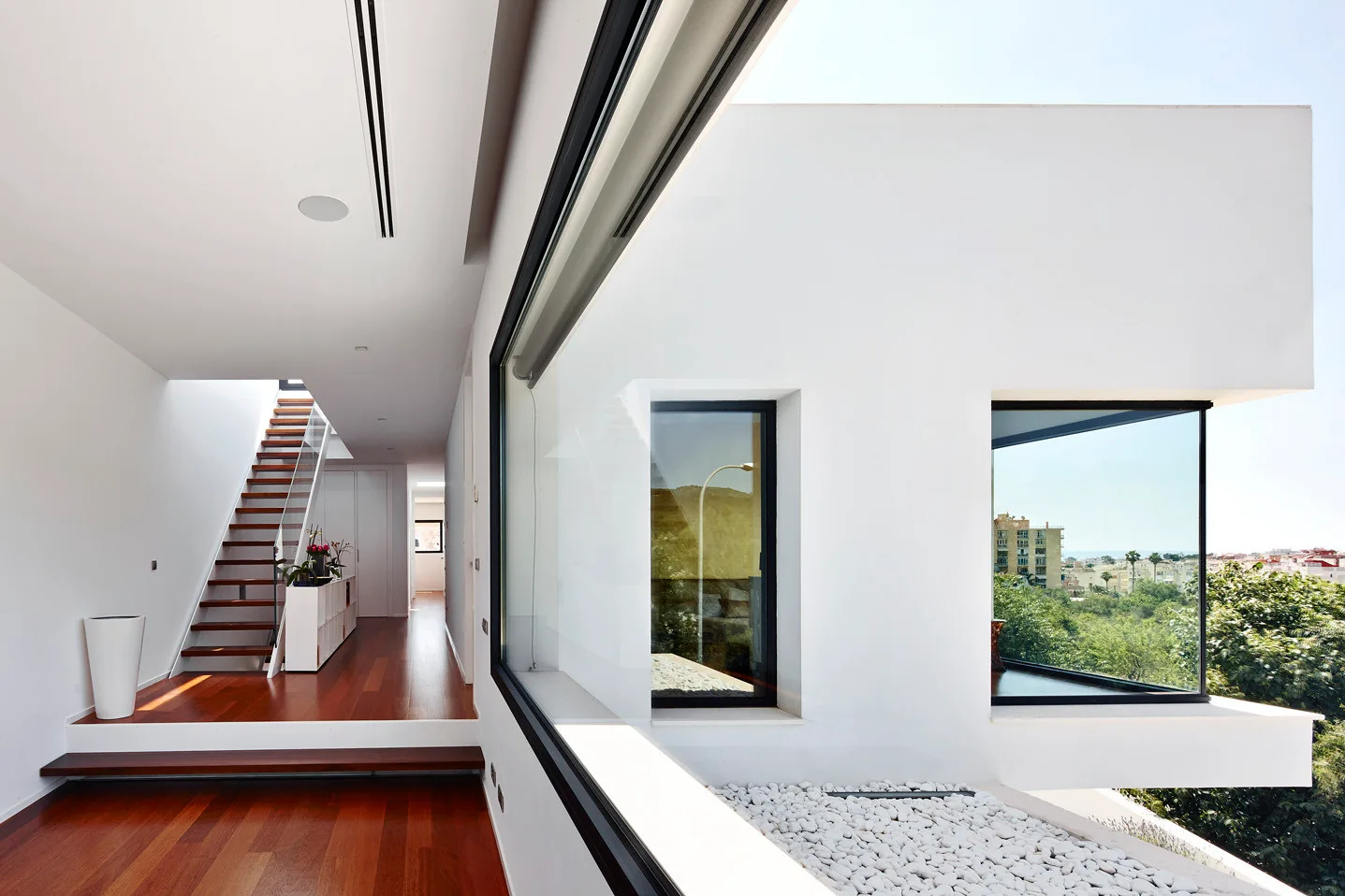 A modern hallway features warm wood floors, a floating staircase with a glass railing, and large windows looking out onto white gravel.