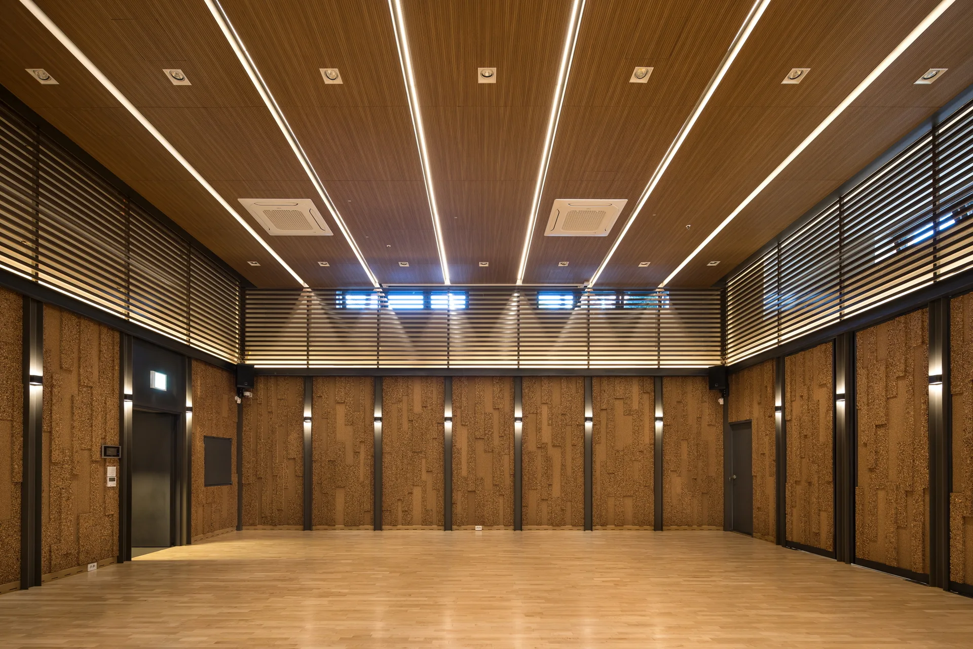 A spacious empty wood-paneled room features textured vertical wall tiles, a horizontal slatted border, and bright linear lights running across the ceiling.