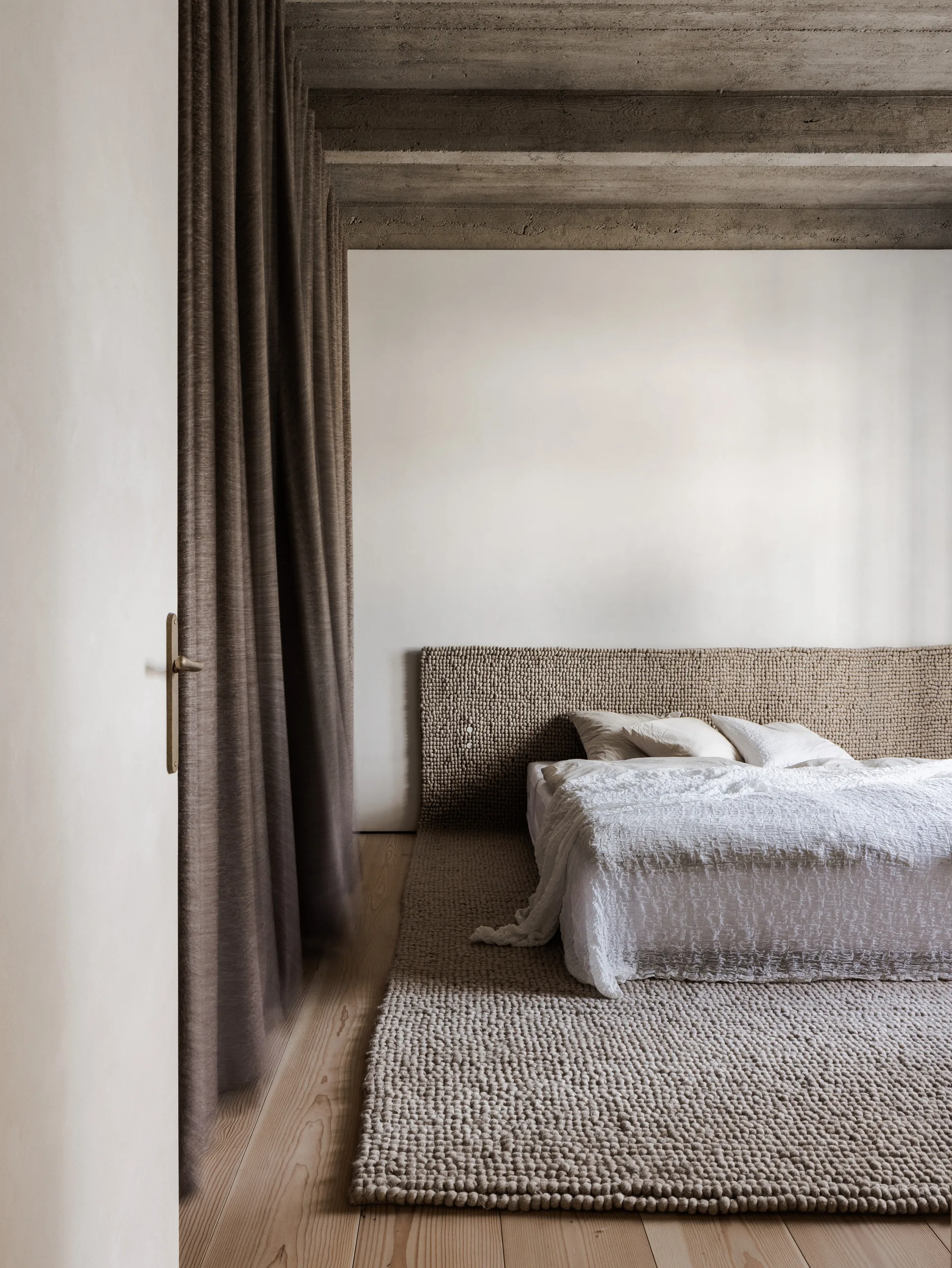 A minimalist bedroom featuring a textured loop rug and matching headboard beneath raw concrete ceiling beams.