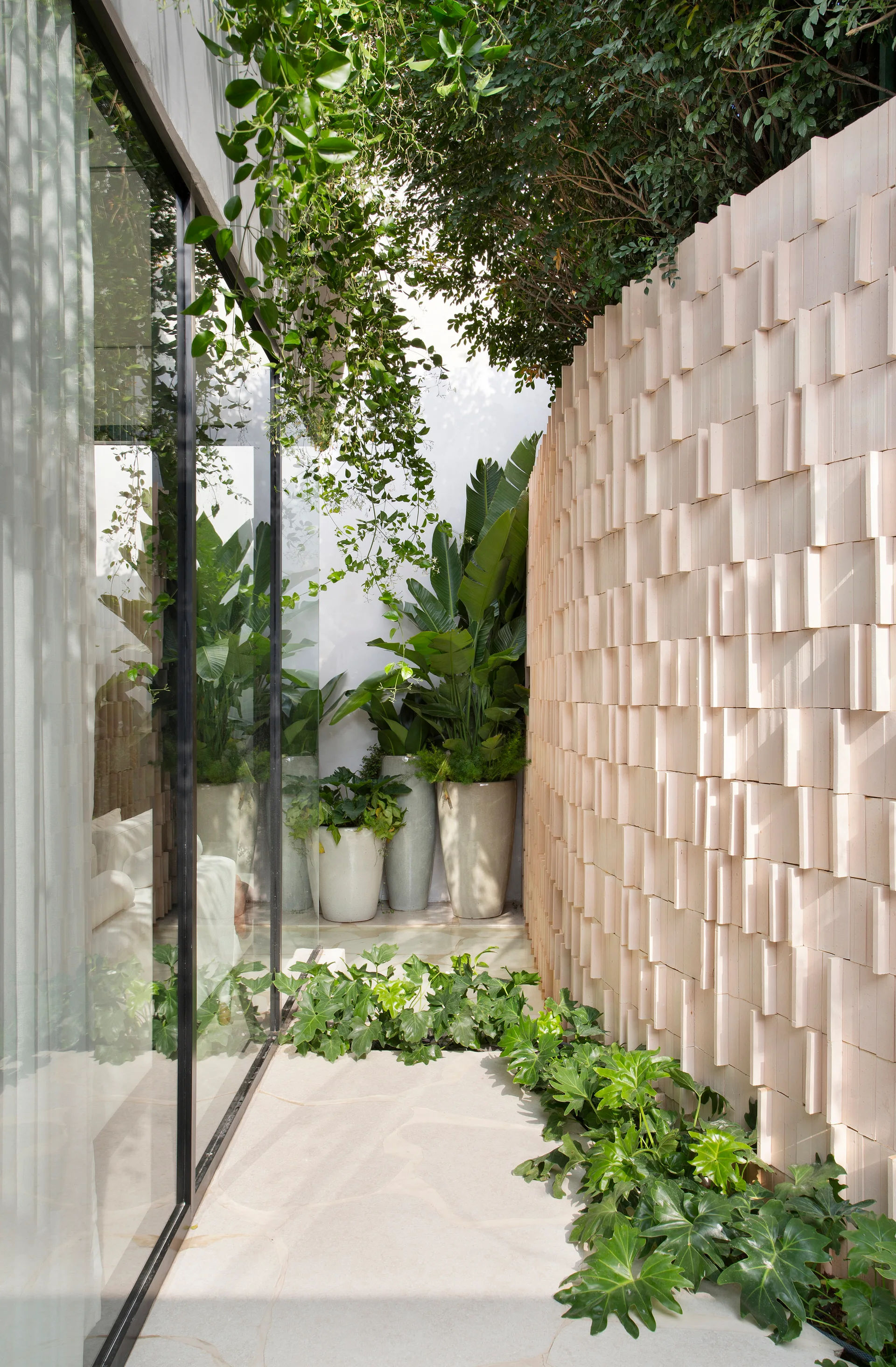 A sunlit outdoor path features a curved blush-pink textured brick wall, stone pavers, potted tropical plants, and a reflective glass window.