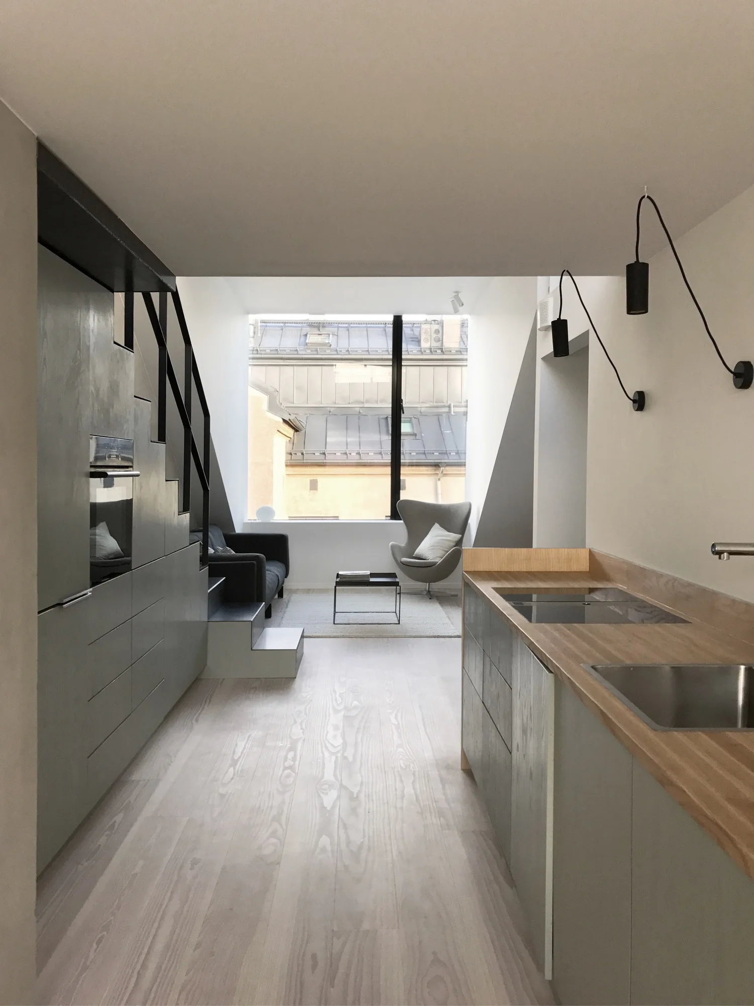 A narrow minimalist kitchen with grey built-in cabinets and integrated stairs leading to a bright living area with a large window.