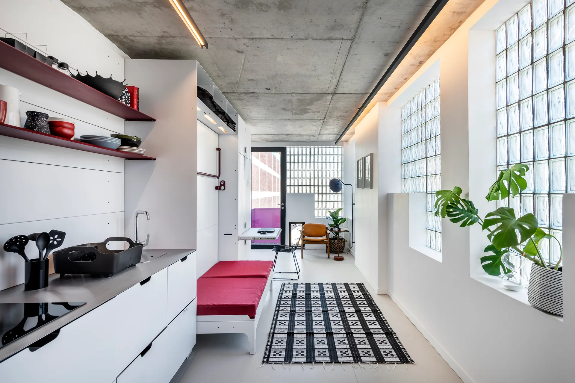 A modern studio apartment kitchen features white cabinetry, a red daybed, a checkered floor rug, and a large glass block window.